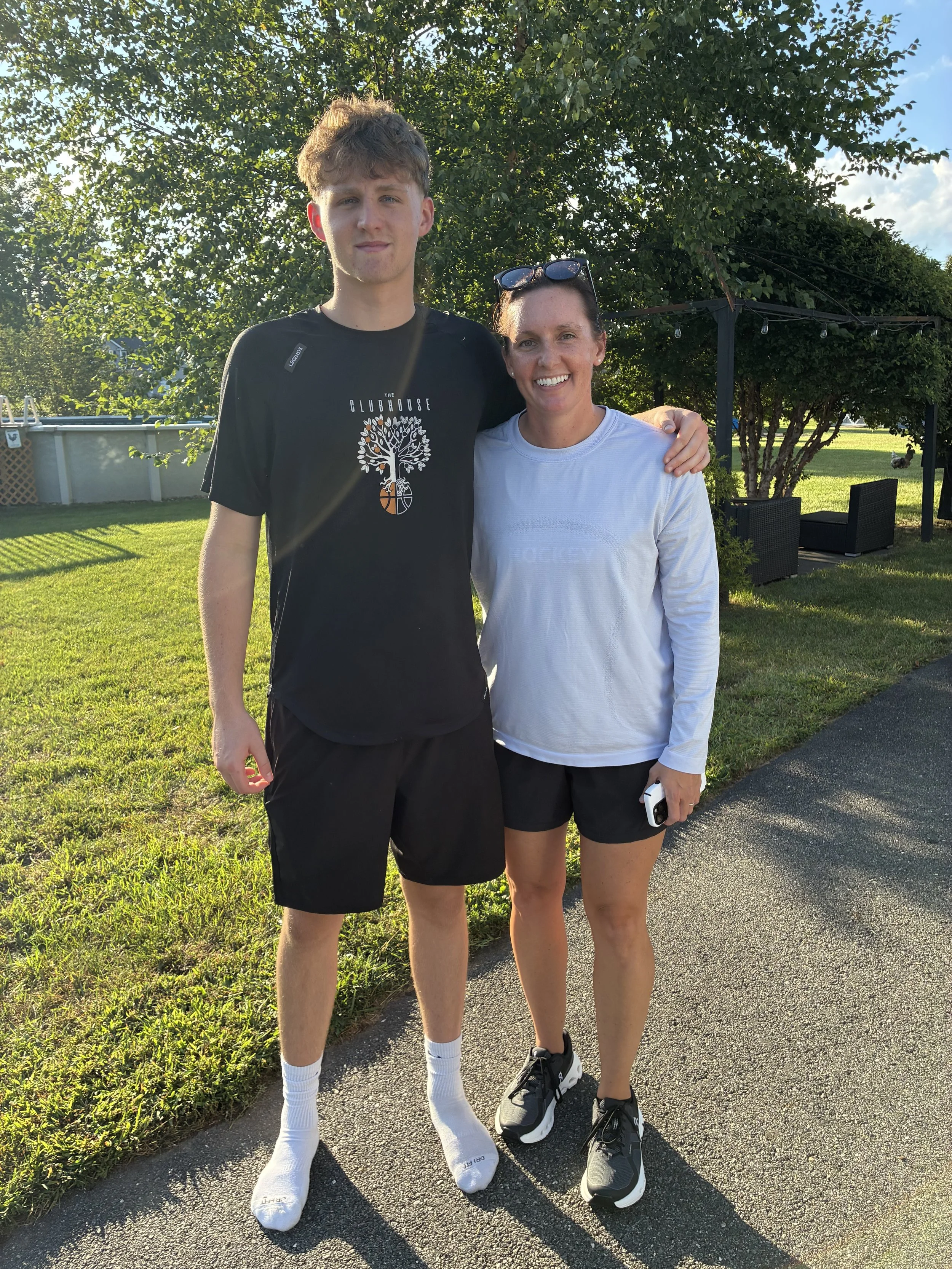 A young man and a woman stand outdoors on a sunny day, smiling and posing together. The young man is wearing a black t-shirt with a graphic design, and the woman is wearing a white long-sleeve shirt and sunglasses on her head. They are standing on a paved path with green grass and trees in the background.