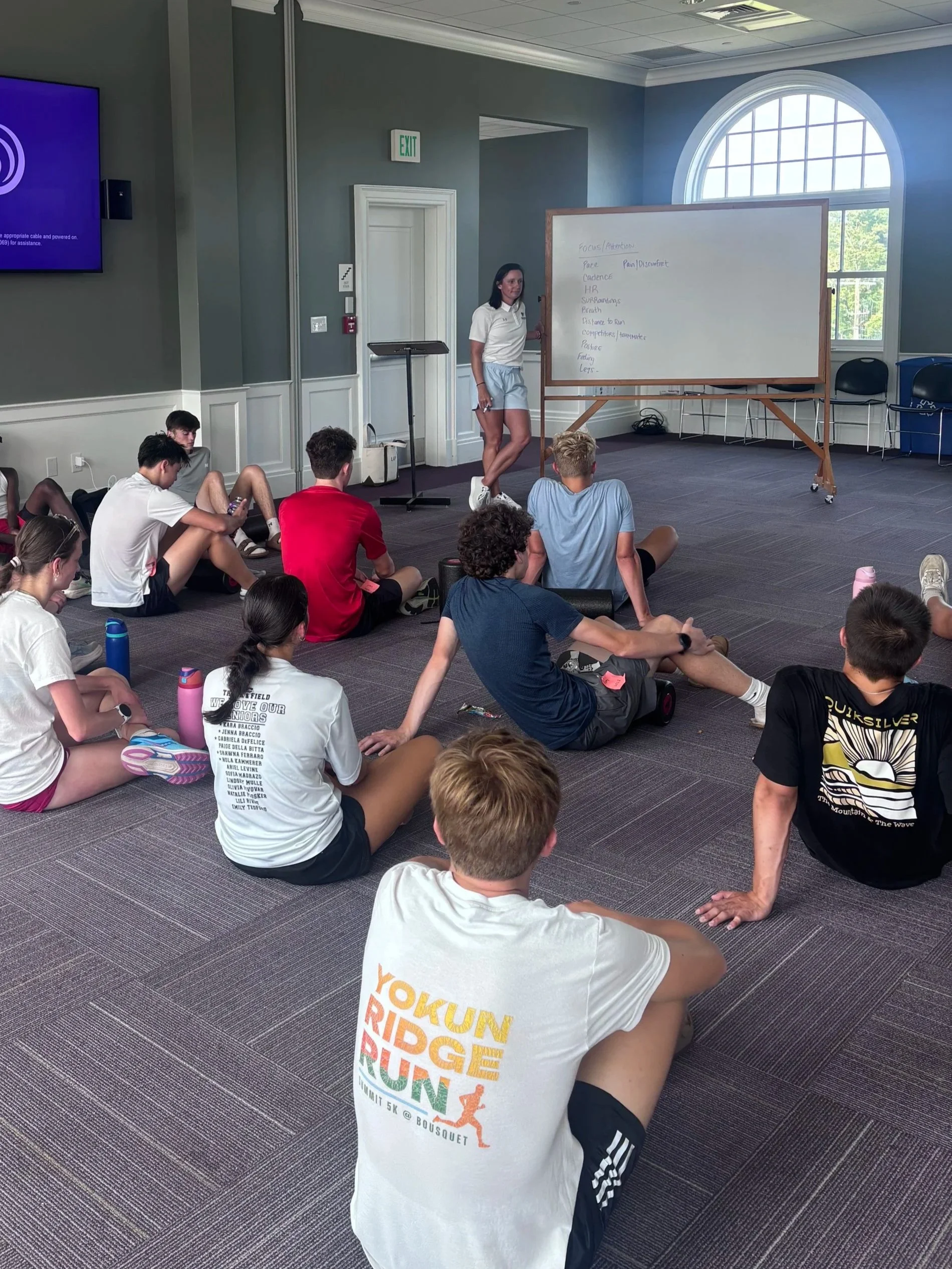 A group of young people sitting on the floor listening to a woman giving a presentation in a room with a large window. The woman is standing beside a whiteboard with notes related to focus and attention. One person in the foreground is wearing a white t-shirt with text promoting a mountain biking event.