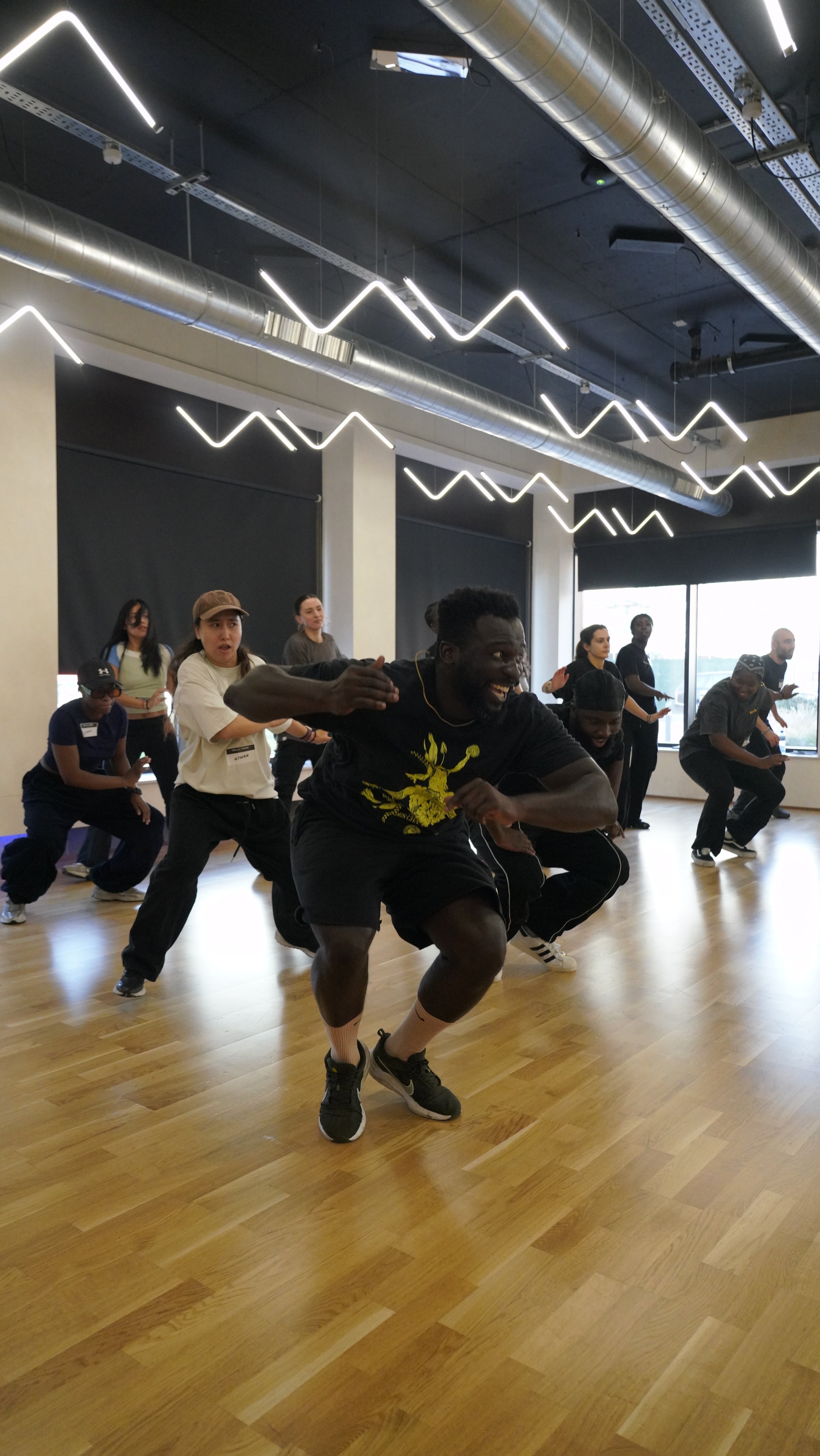 A group of people participating in a dance or fitness class in a studio with wooden floors, large windows, and modern lighting fixtures.