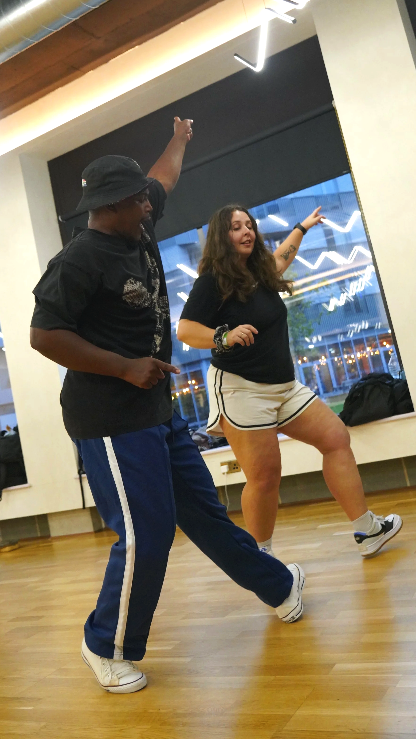 Two people dancing indoors in front of a large mirror, with city lights visible through the window behind them.