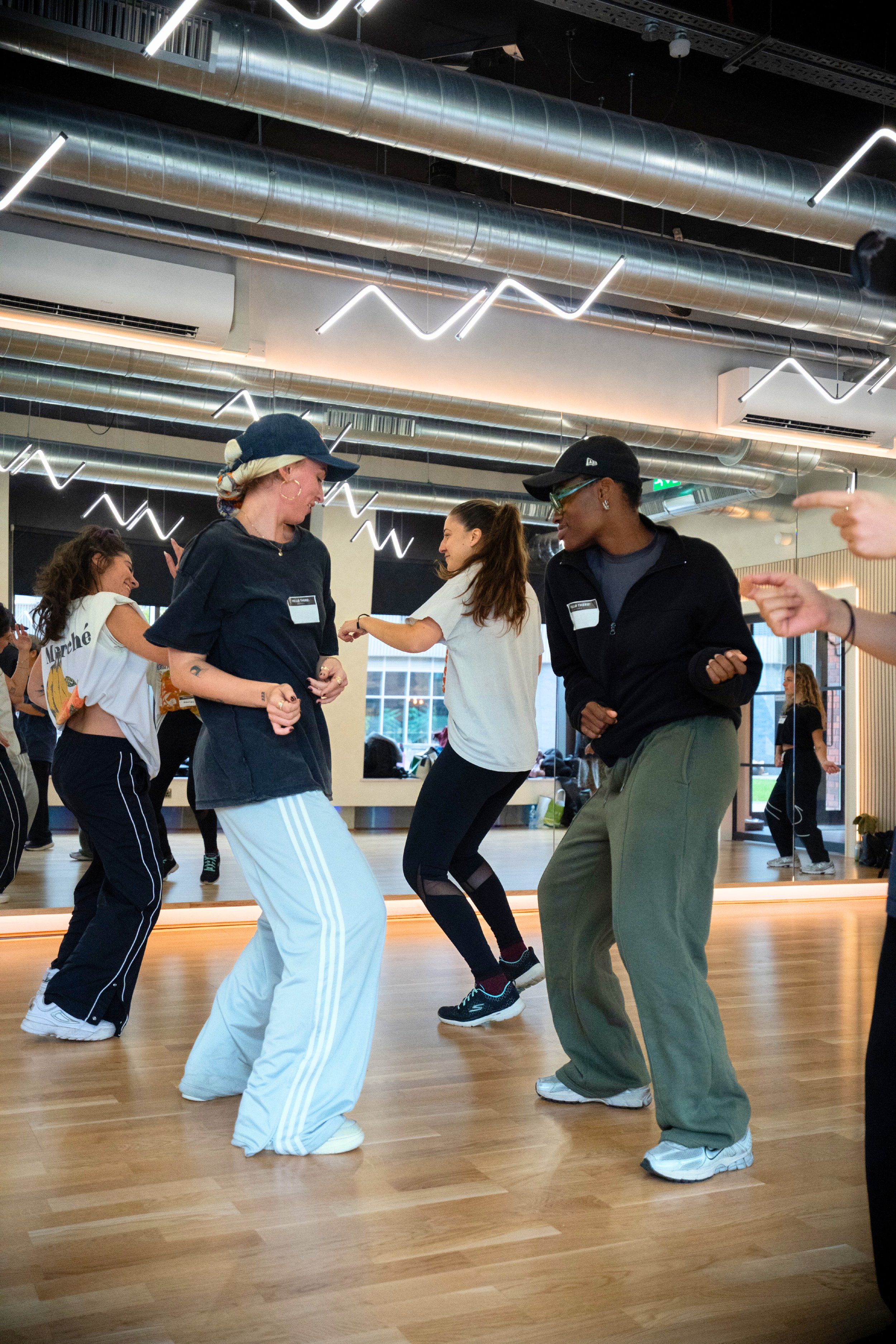 People dancing in a modern dance studio with wood floors, large mirrors, and industrial ceiling with metal pipes and neon light designs.