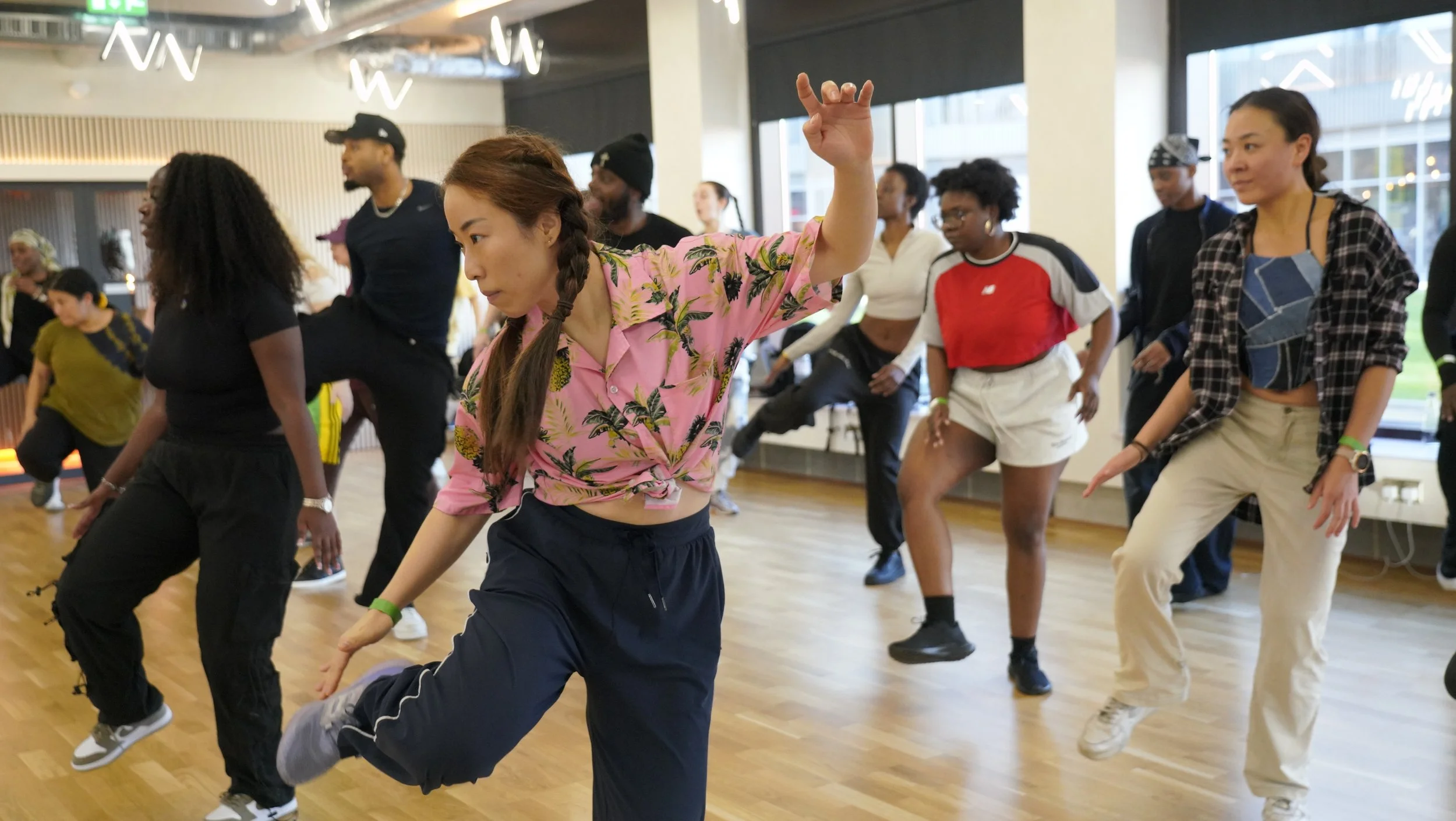 A diverse group of people dancing in a studio with wooden floors and large windows, led by a woman in a pink floral shirt, wearing black pants, with others in casual athletic wear following her lead.