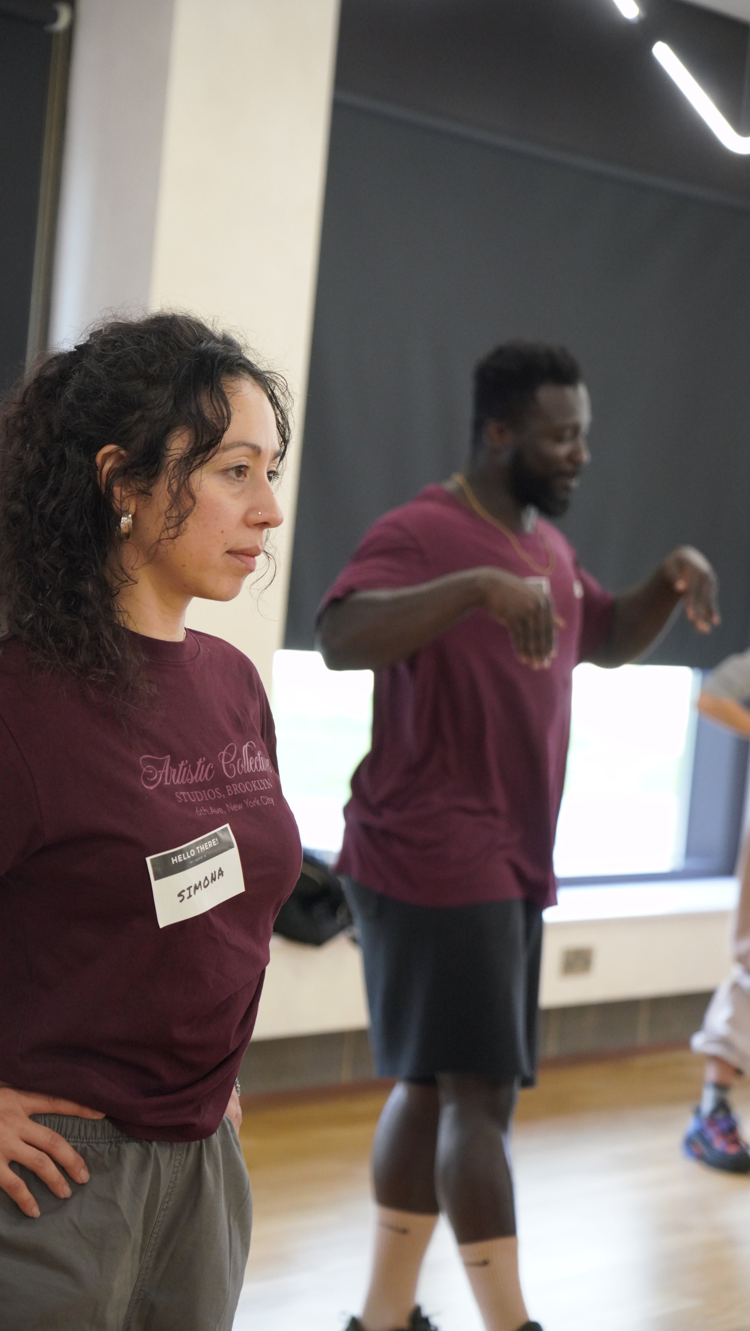 A woman with curly brown hair, wearing a maroon t-shirt labeled "Simona" and gray pants, standing with her hands on her hips in a room with a black wall and window, while a man in a maroon t-shirt and black shorts dances or moves with his arms raised