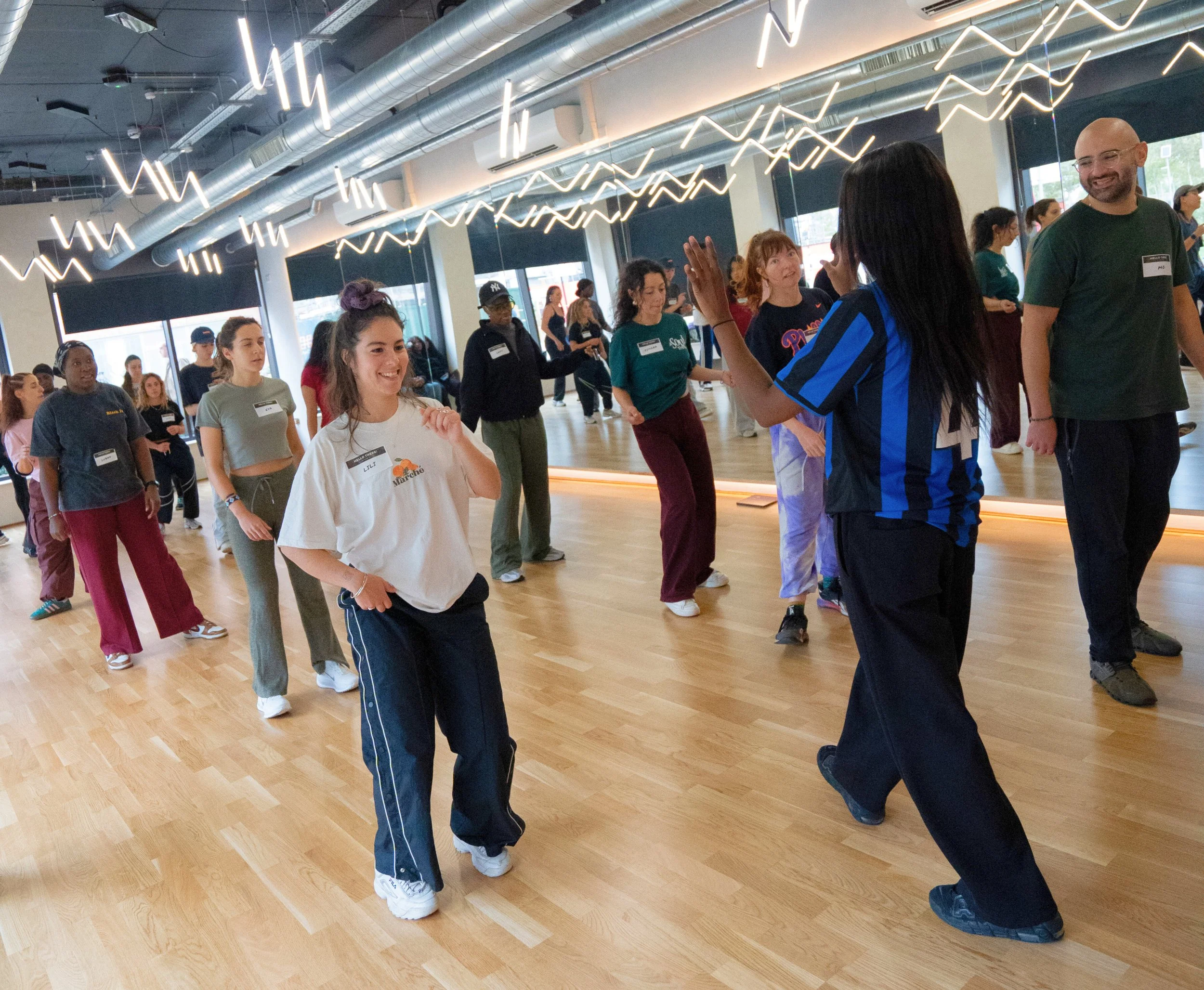 A group of people dancing in a studio with wooden floors, large windows, and modern lighting fixtures.