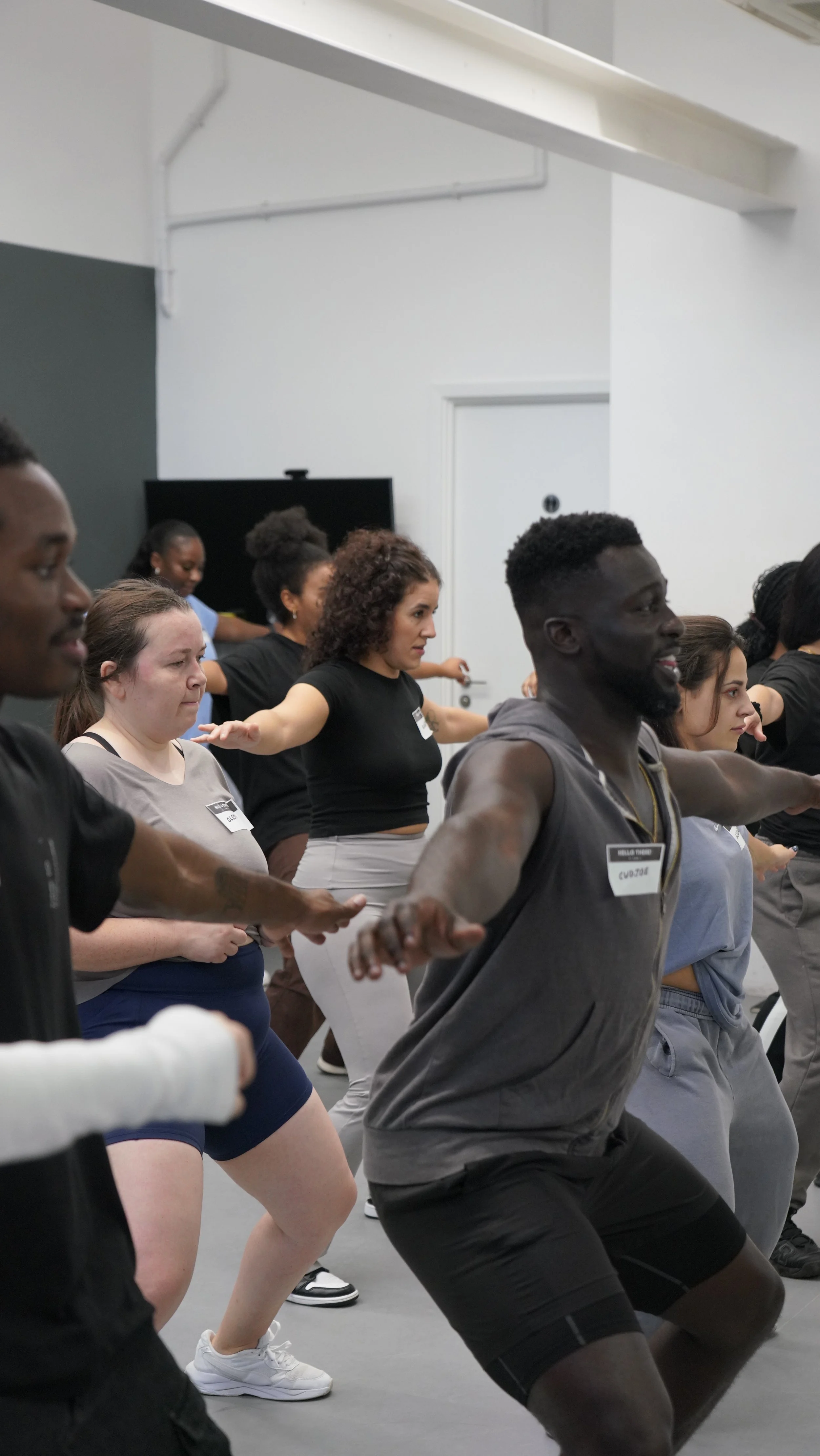 Group of diverse people participating in a fitness or dance class, standing with arms extended to the sides, in a studio with white walls.
