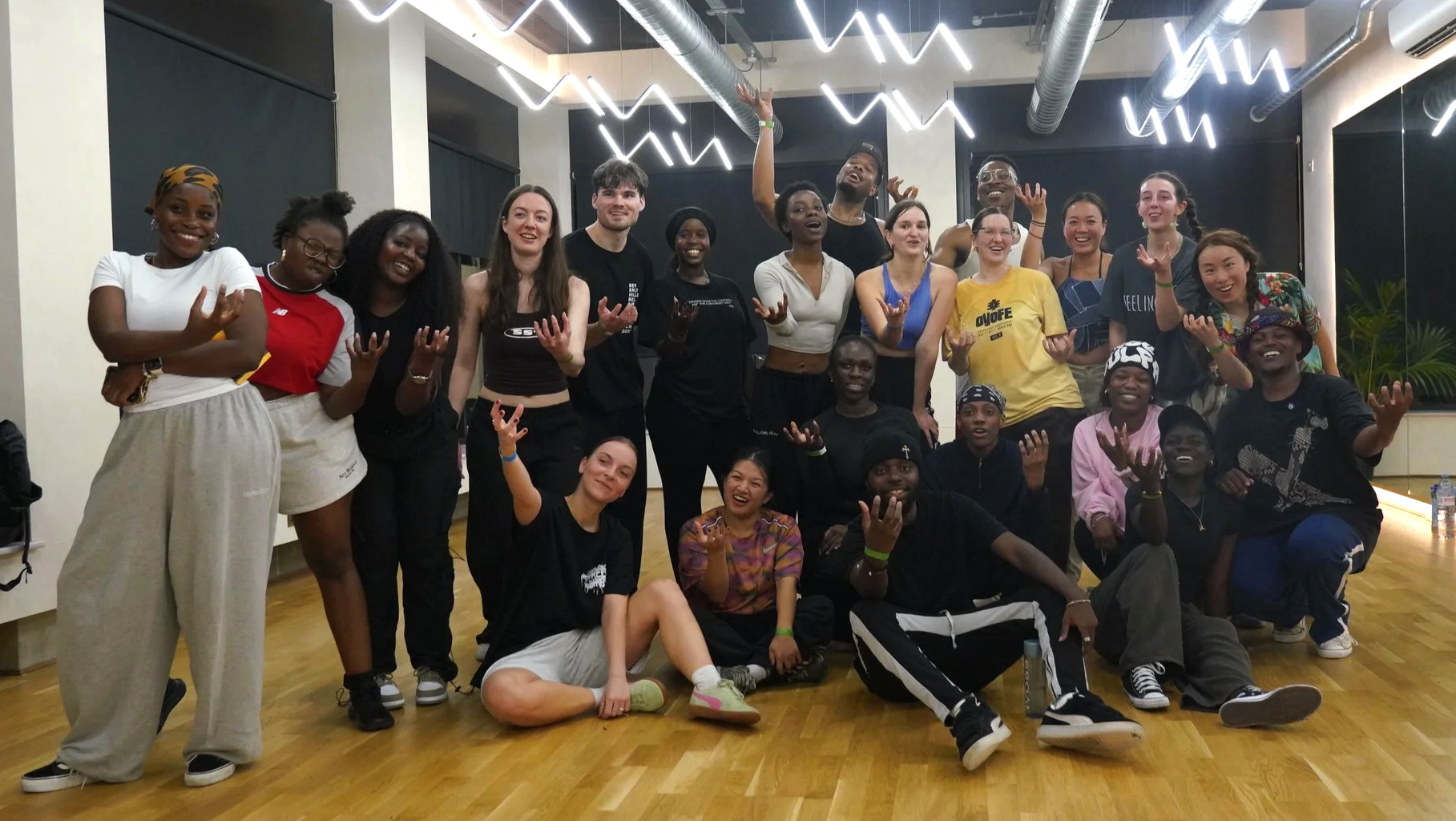 Group of diverse young adults smiling and making hand gestures indoors with modern ceiling lights.