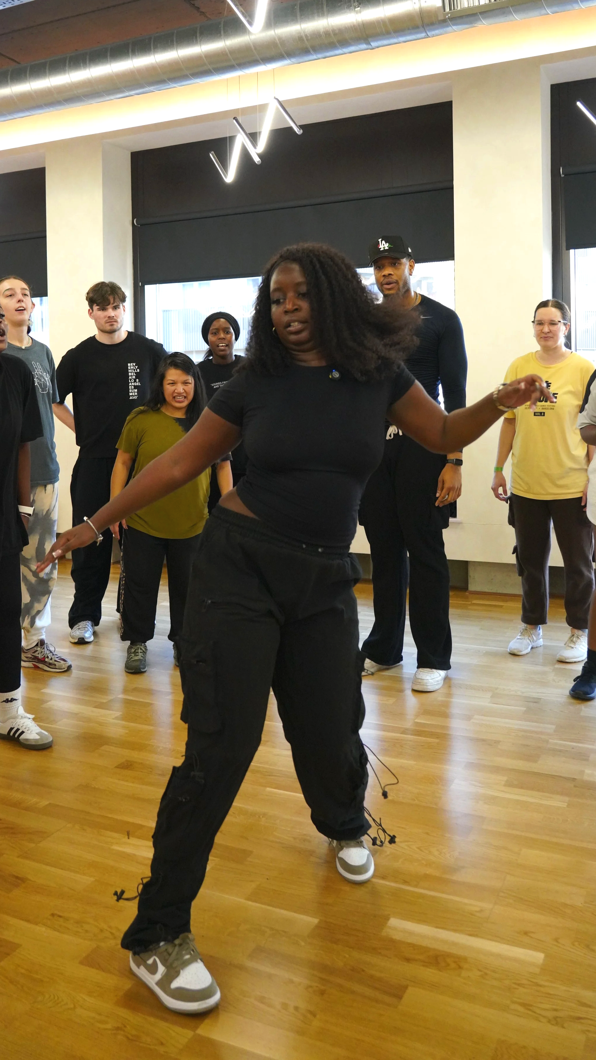 Group of diverse people in a dance studio, with one woman leading a dance move in the foreground.