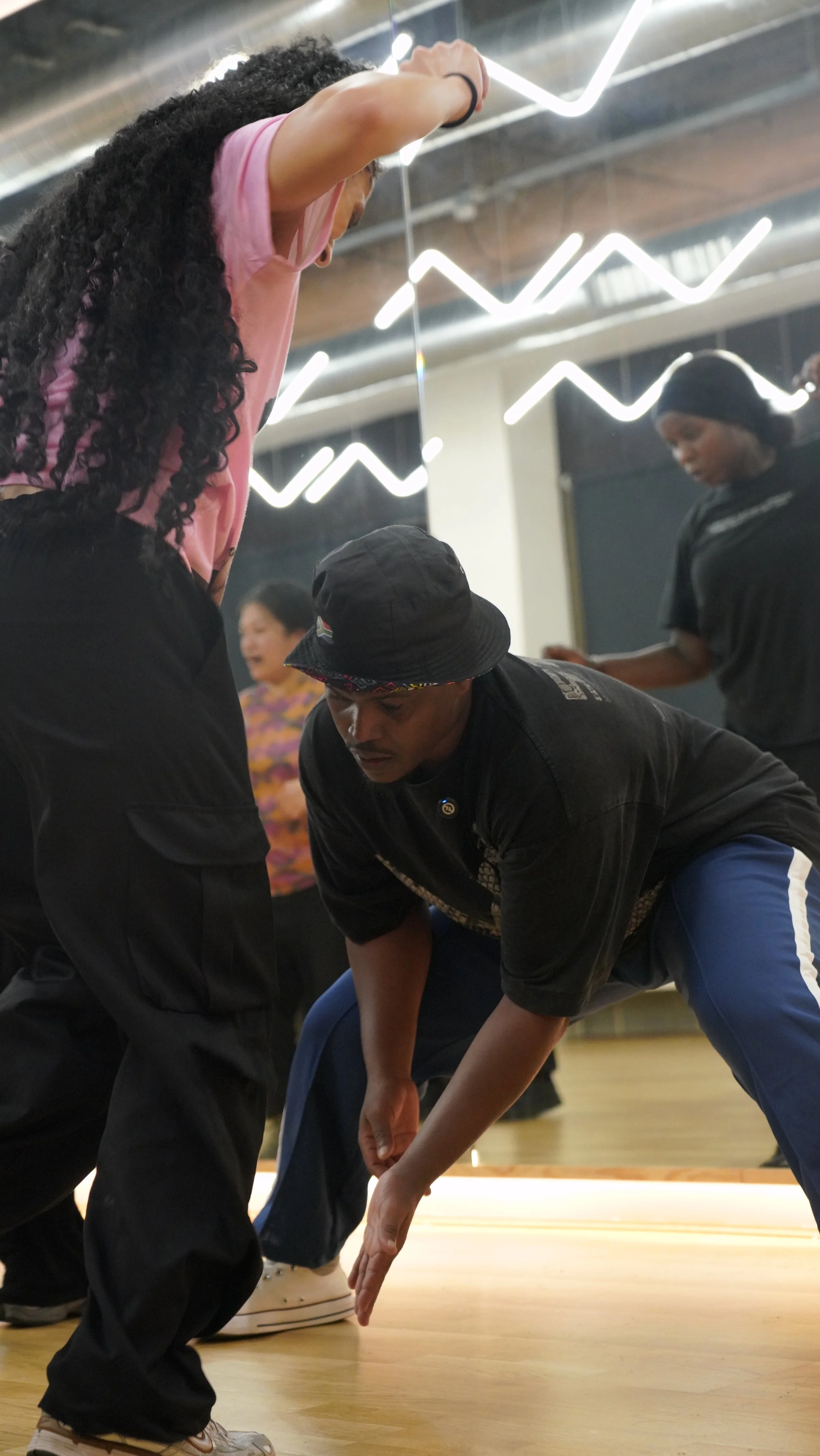 A dance instructor demonstrates a move to a group of dancers in a studio with neon light decorations on the ceiling.
