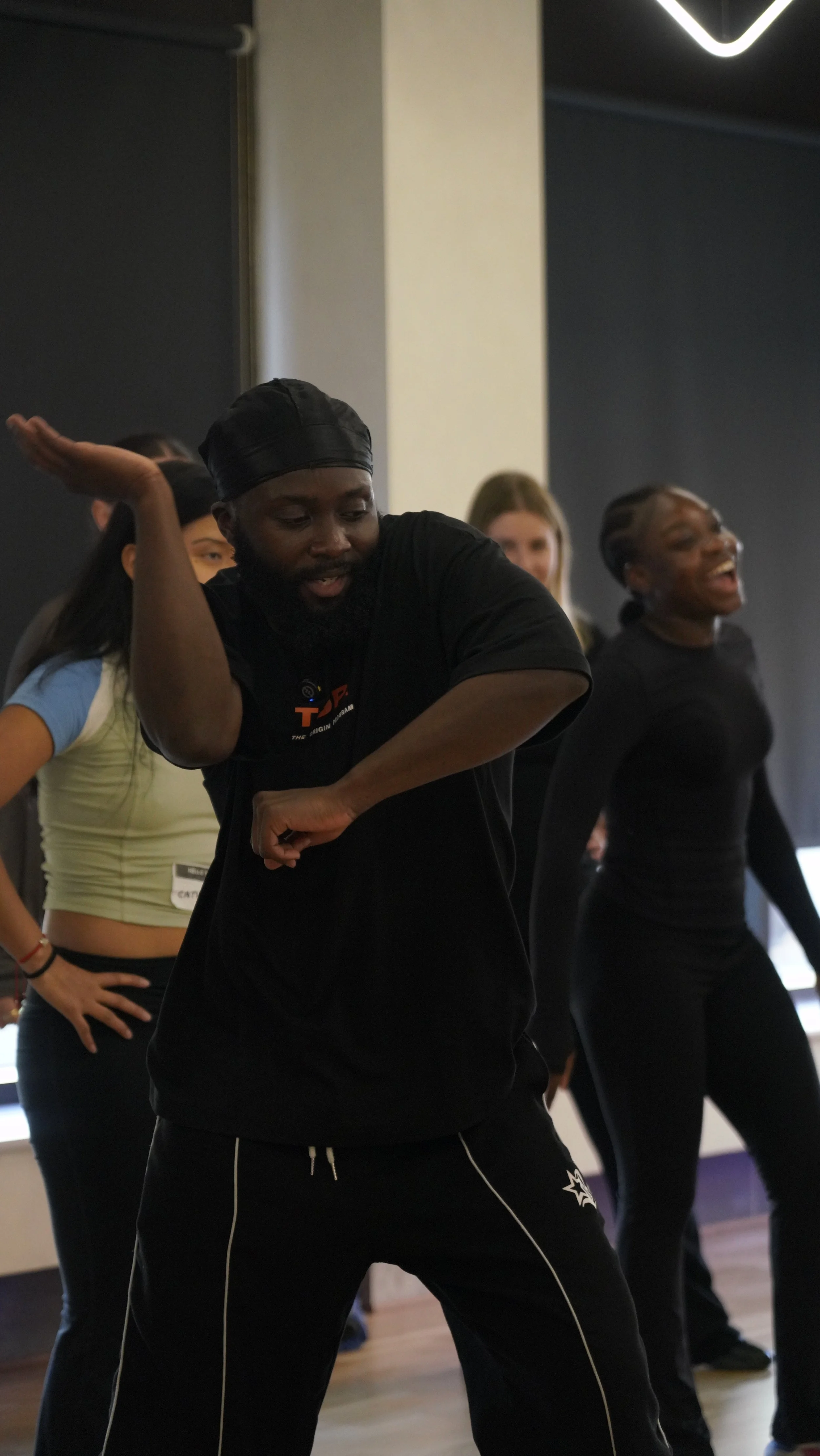 A group of people dancing in a studio, with a man in the foreground wearing a black head wrap and black clothing, smiling, and others in the background also dancing and smiling.