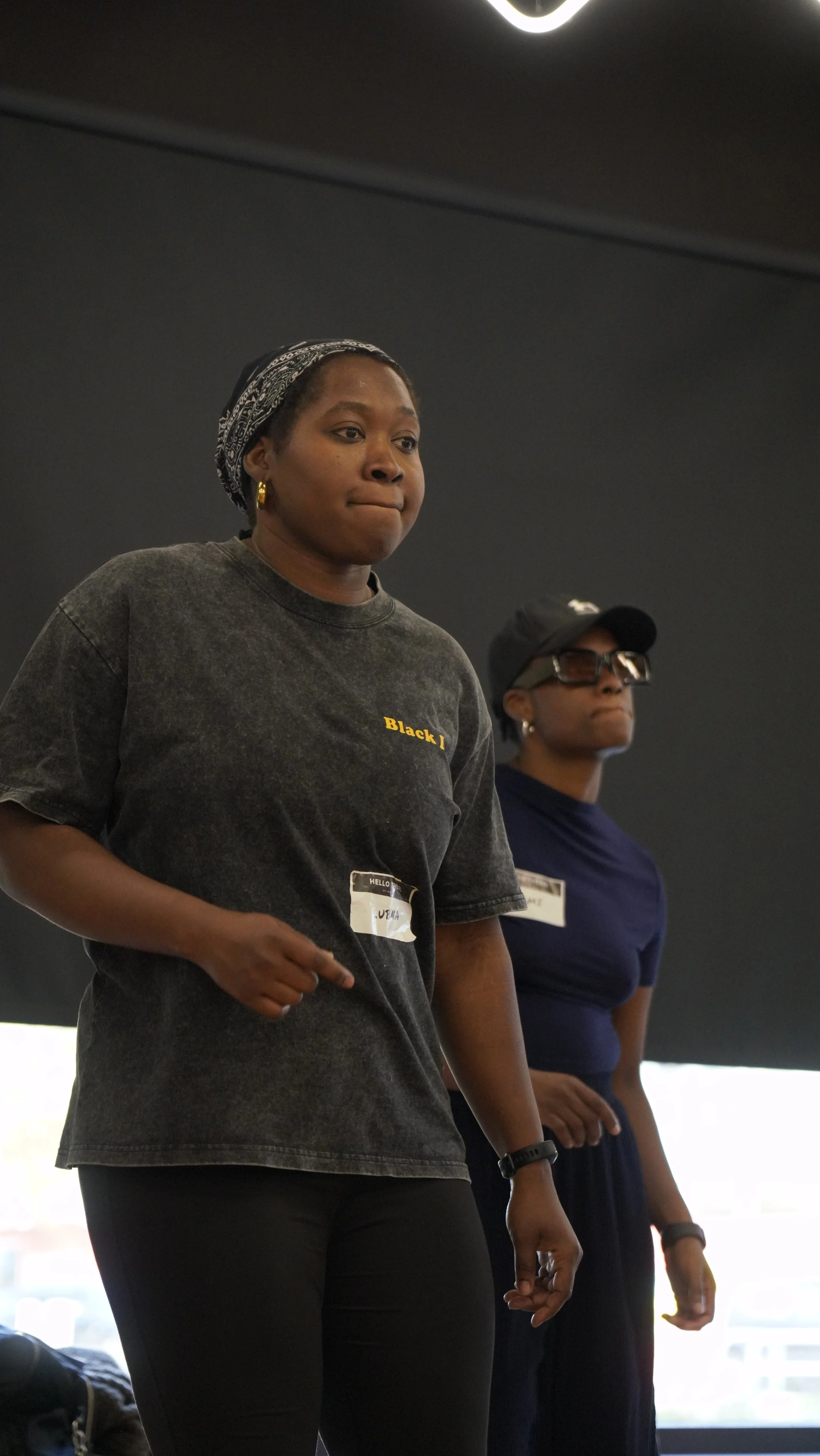 Two women standing indoors, one in the foreground wearing a black T-shirt with yellow text and a name tag, the other in the background wearing a dark blue shirt, glasses, and a black cap, both looking attentive.