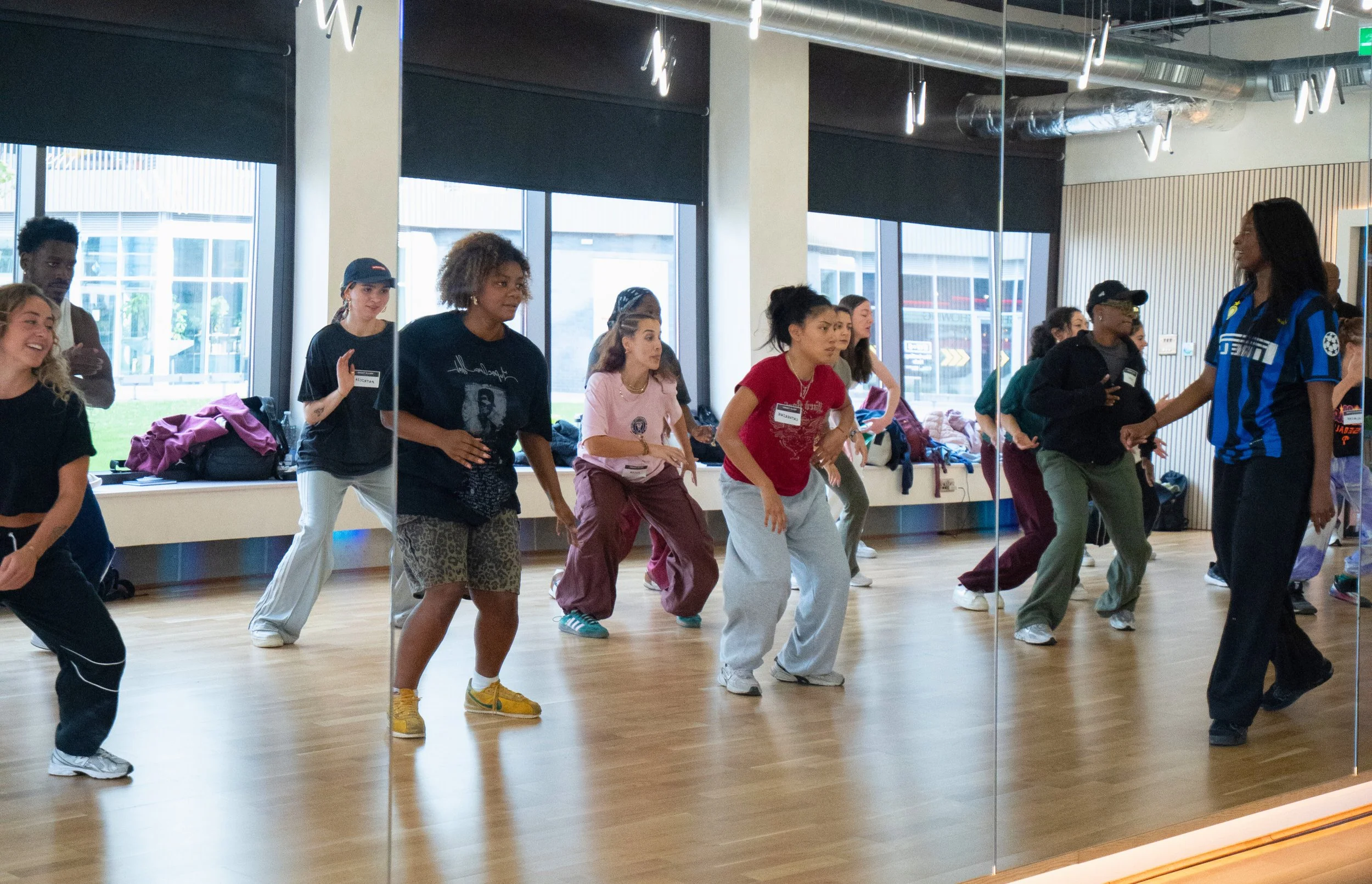A group of diverse women and girls participating in a dance or fitness class in a studio with wood floors, large windows, and a mirrored wall.
