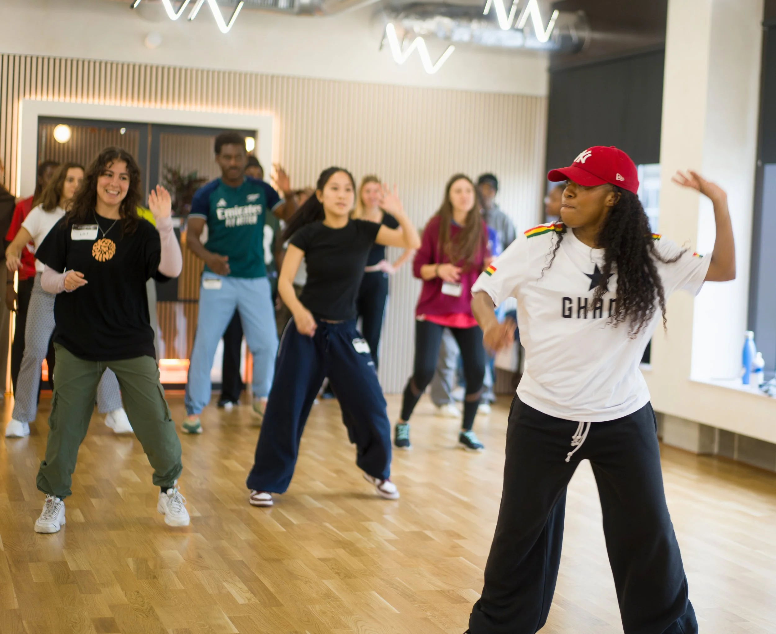 A group of diverse women participating in a dance class or dance workshop in a studio with wooden floors and modern interior lighting. The instructor or lead dancer is a woman wearing a white T-shirt with black pants and a red cap, demonstrating danc