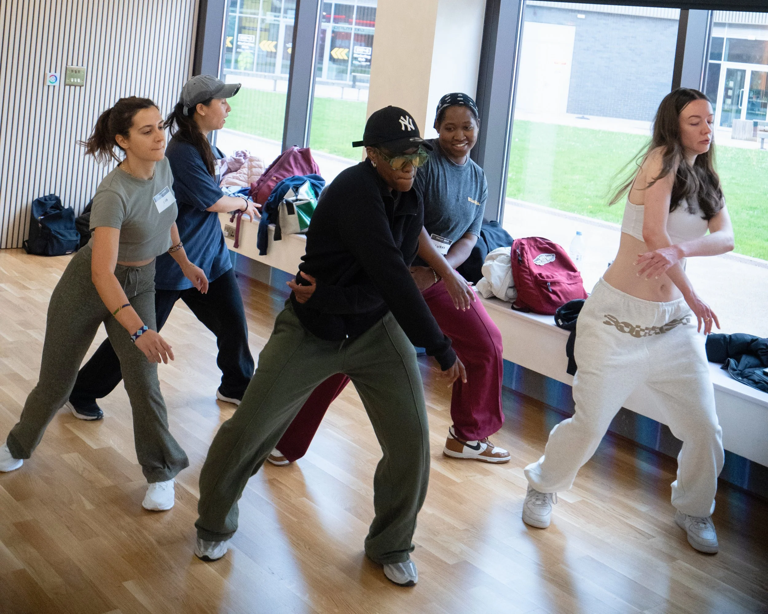 Group of five women dancing together in a room with large windows, with backpacks and personal items on the windowsill.