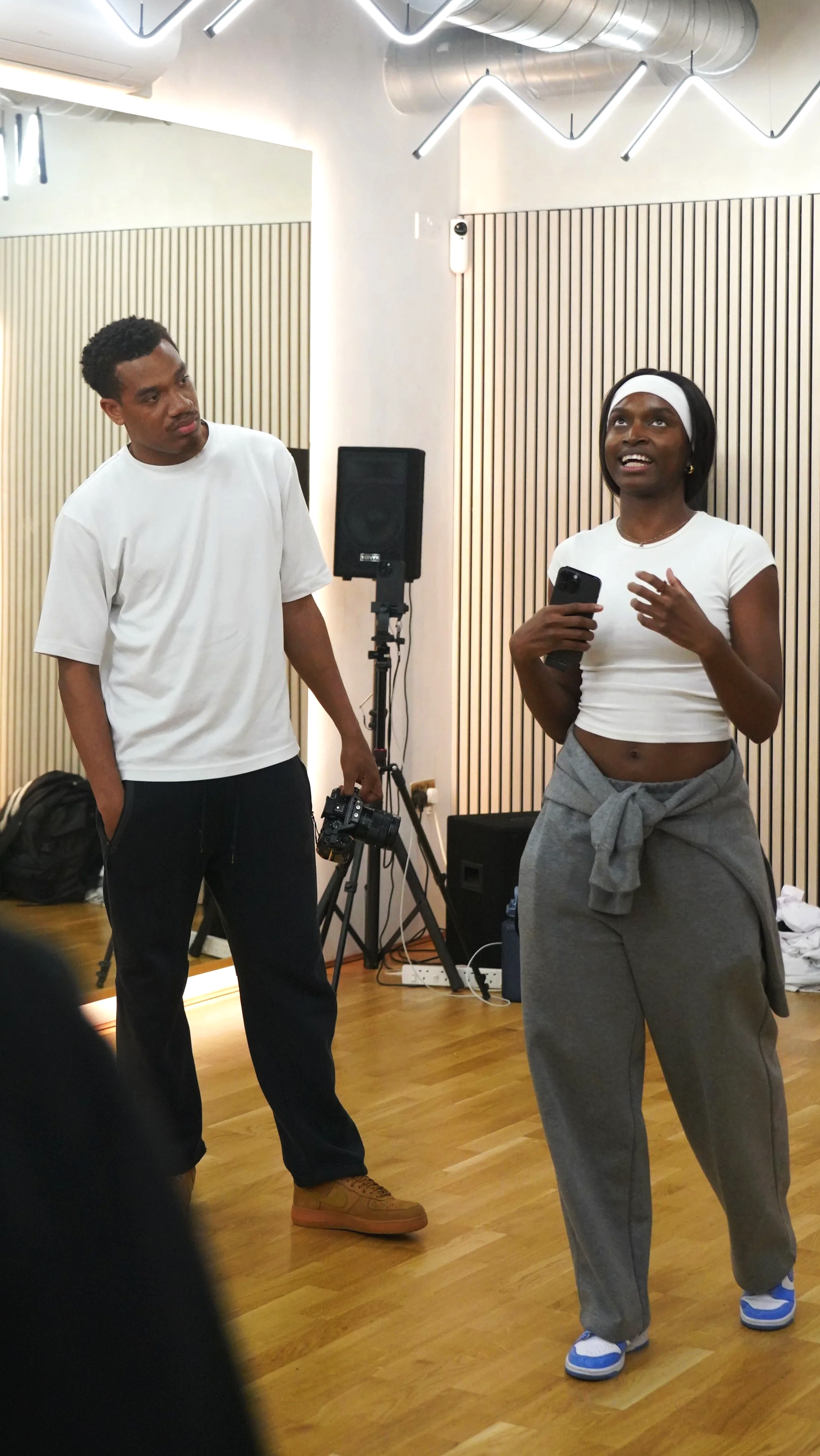 Two people in a dance studio or rehearsal space, one man and one woman. The woman is talking and holding a phone, wearing workout clothes and a headband. The man is holding a camera, listening attentively. The room has wood flooring, sound panels on the wall, and professional lighting fixtures on the ceiling.