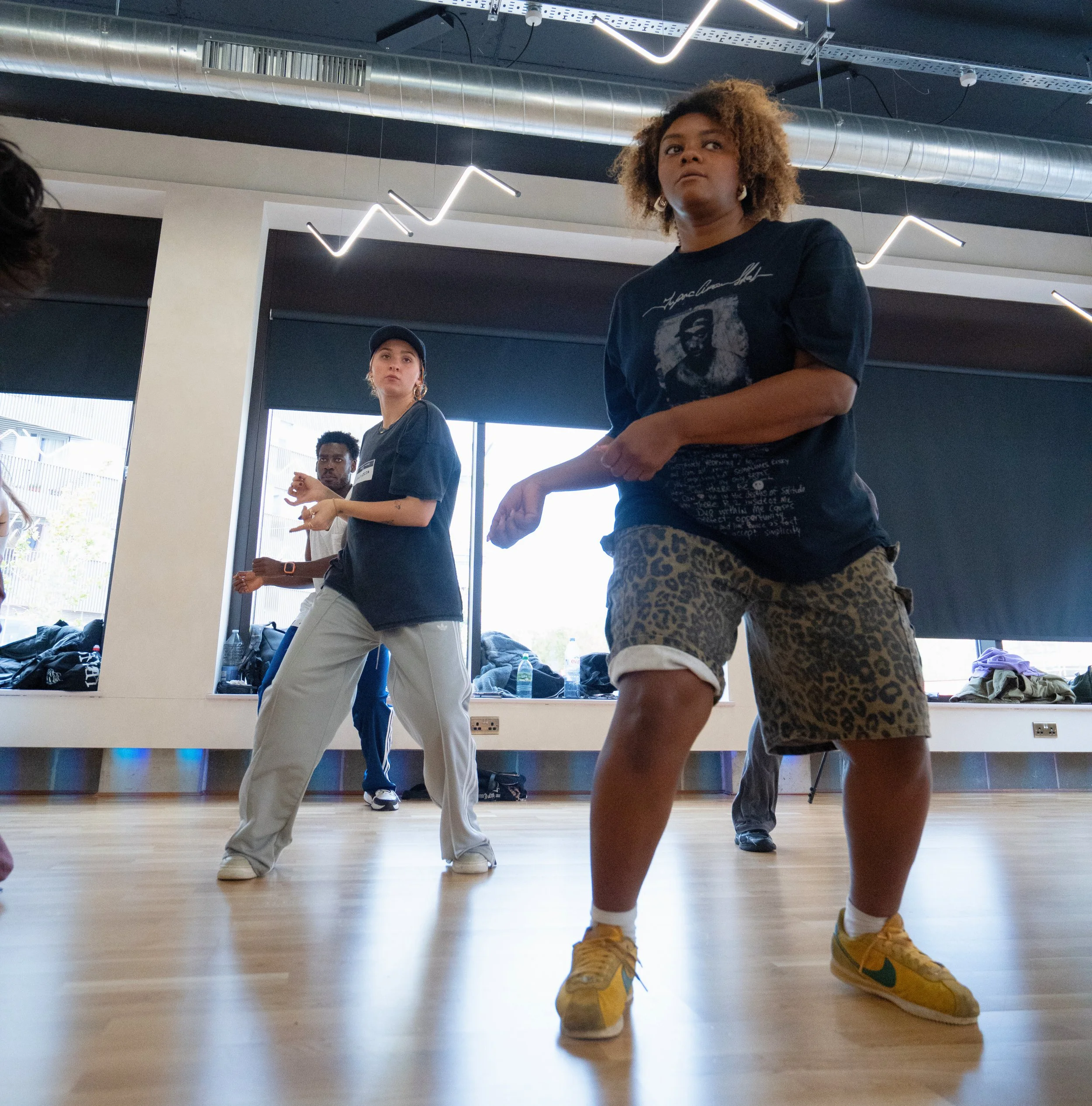 Three people dancing in a studio with a wooden floor and large windows, with LED light fixtures and ventilation pipes on the ceiling.