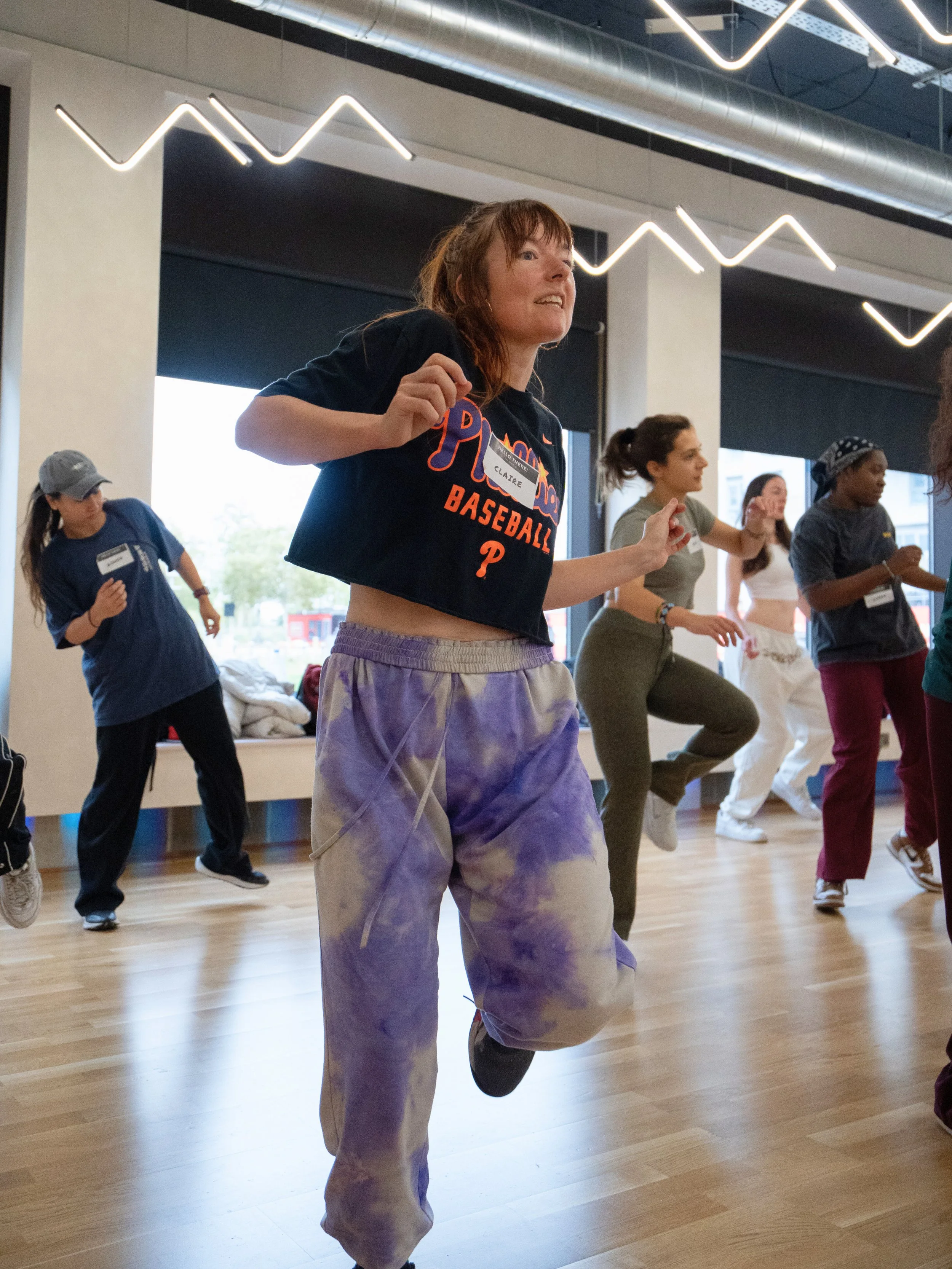 Group of women exercising indoors, leading with a woman in a black cropped T-shirt and purple tie-dye sweatpants.