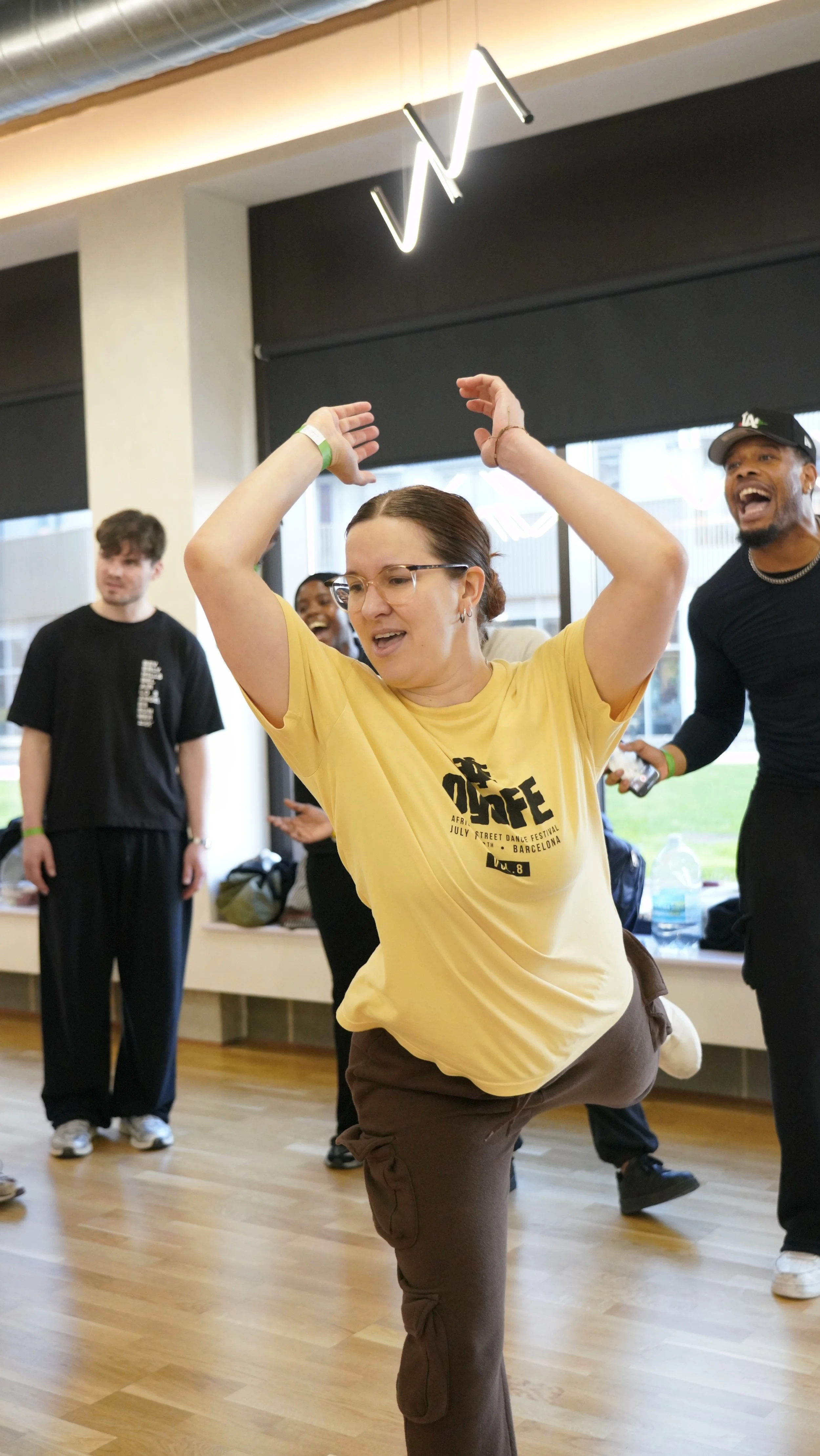 A group of people dance energetically indoors at a dance festival, with a woman in a yellow t-shirt in the foreground raising her arms and lifting her leg, smiling and enjoying the moment.