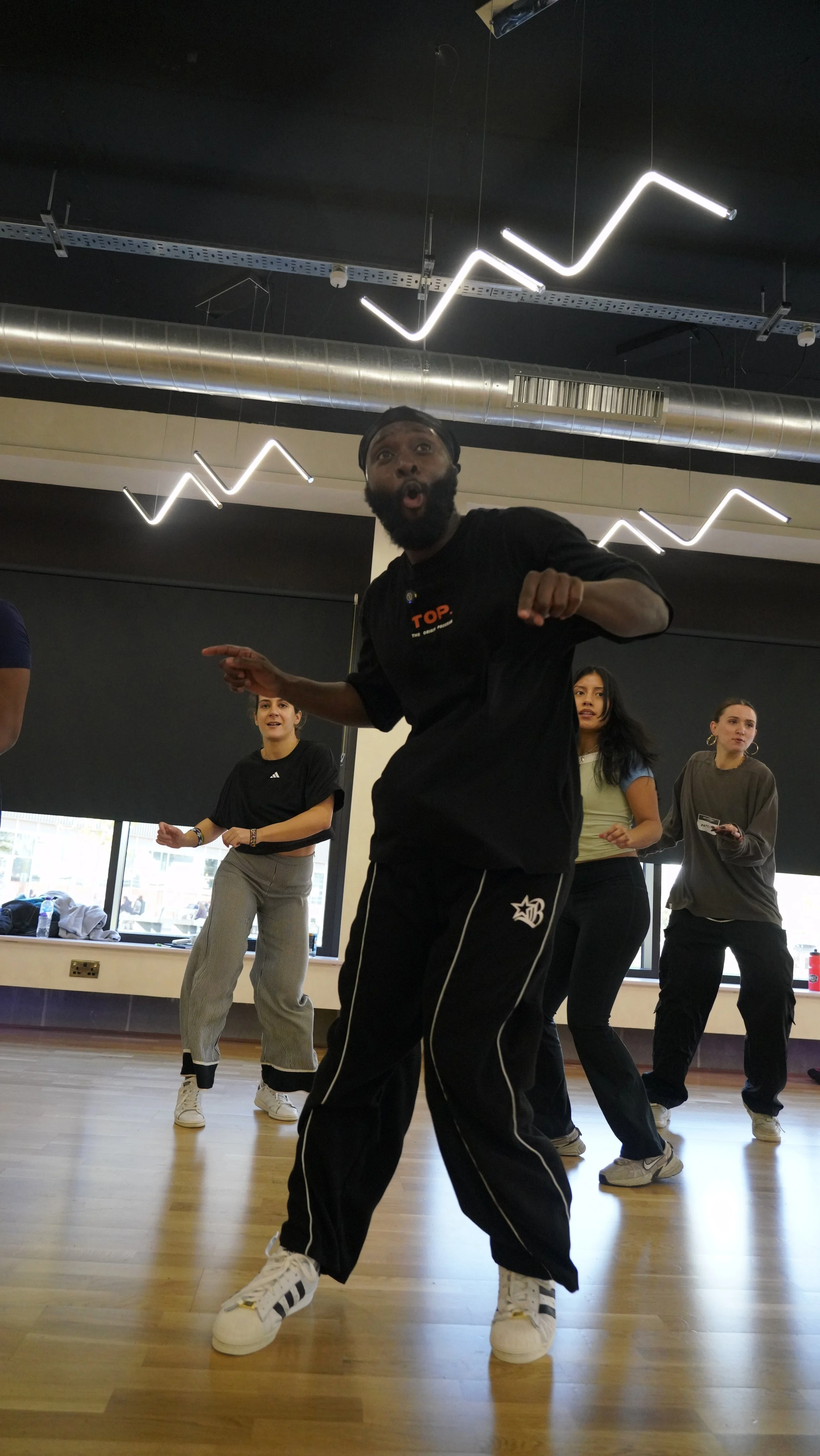 A group of people participating in a dance or fitness class indoors, with a male instructor in the front leading the session, under modern lighting fixtures with zigzag patterns, and large windows in the background.