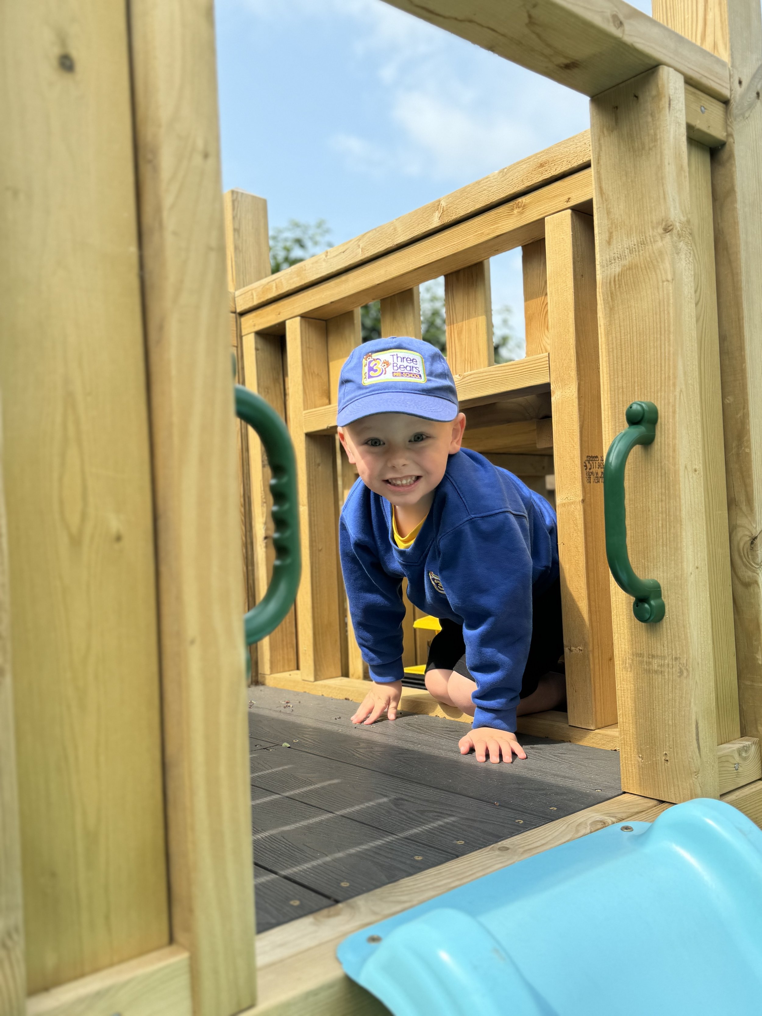 Child crawling out of a wooden playhouse, smiling, wearing a blue cap and blue sweatshirt, on an outdoor playground.