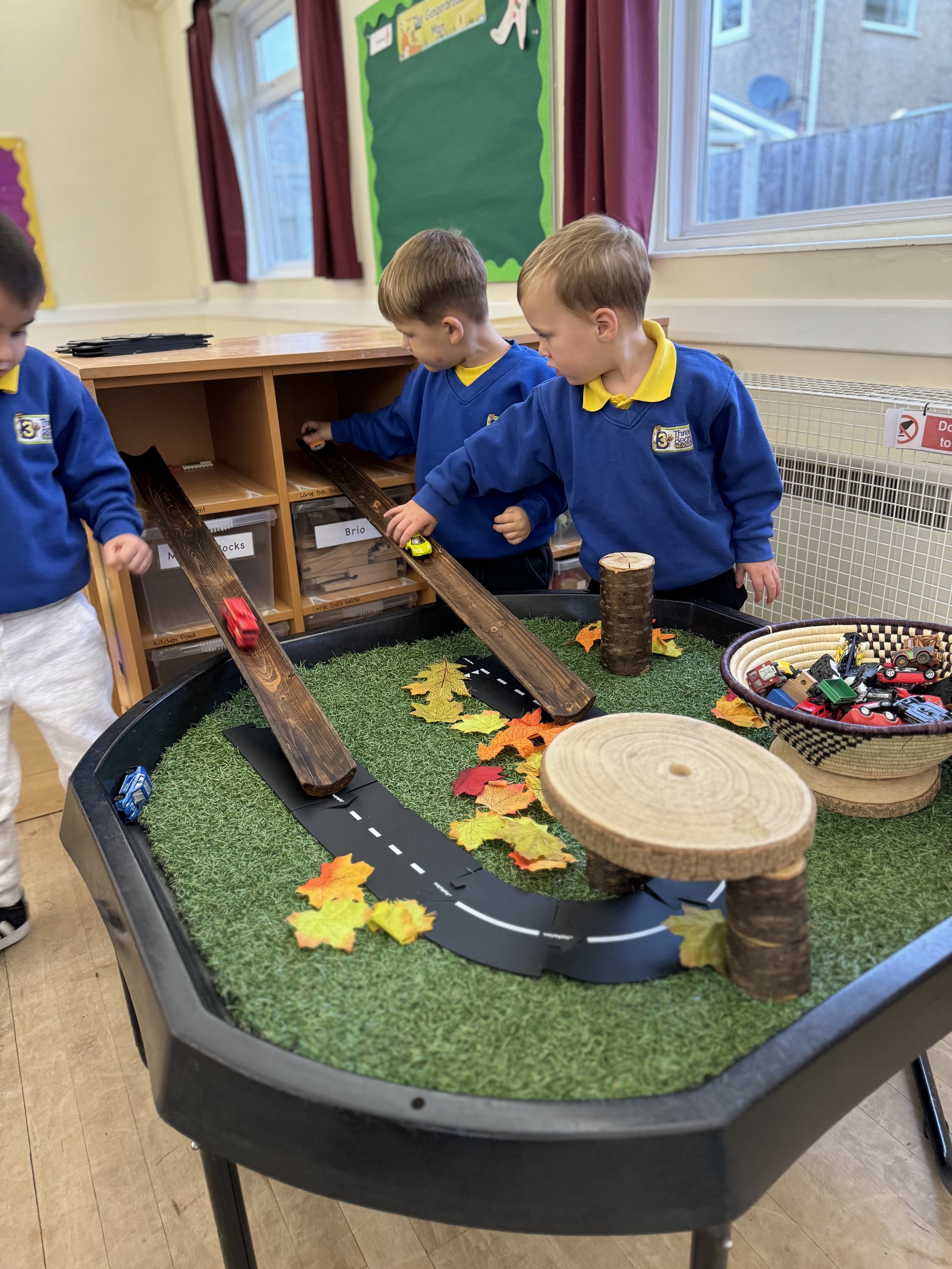 Three young children in blue school uniforms playing with toy cars on a miniature road table with grass, fallen leaves, and wooden logs. The table is in a classroom.