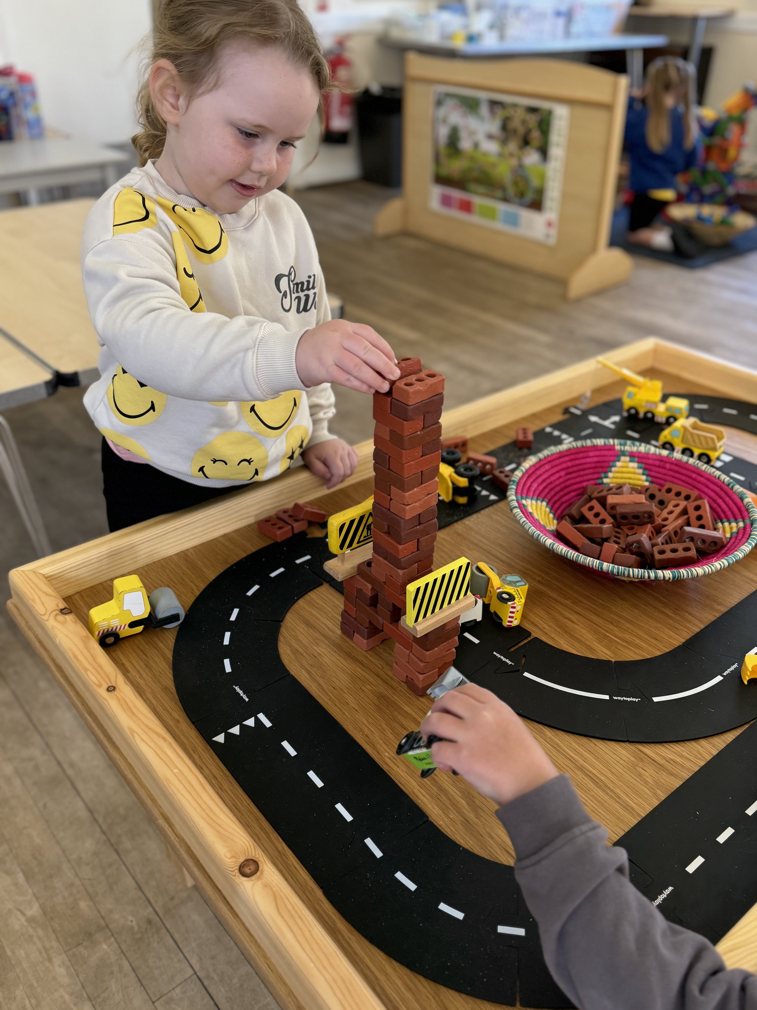 A young girl in a cream-colored sweatshirt with yellow smiley face designs stacking small red bricks into a tall tower on a play roadway table. There is a basket of additional red bricks, small toy construction vehicles, and traffic barriers on the t