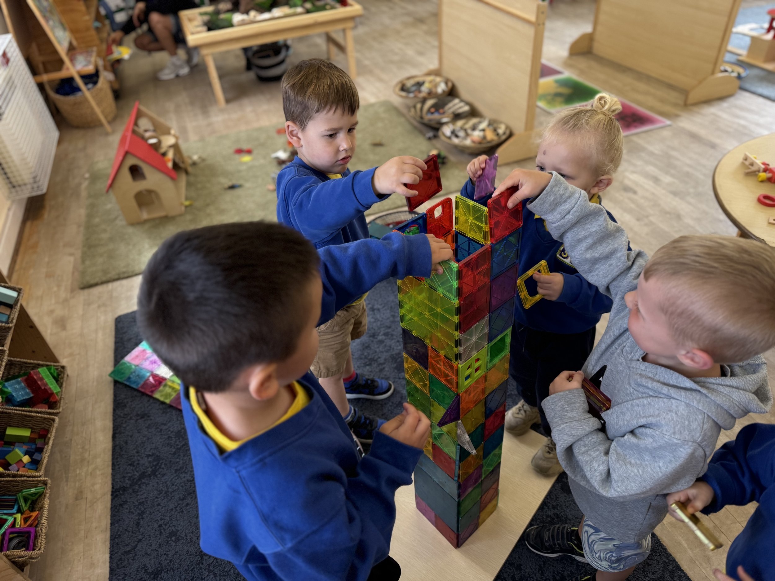 Four children playing with colorful magnetic tiles to build a tower in a classroom.