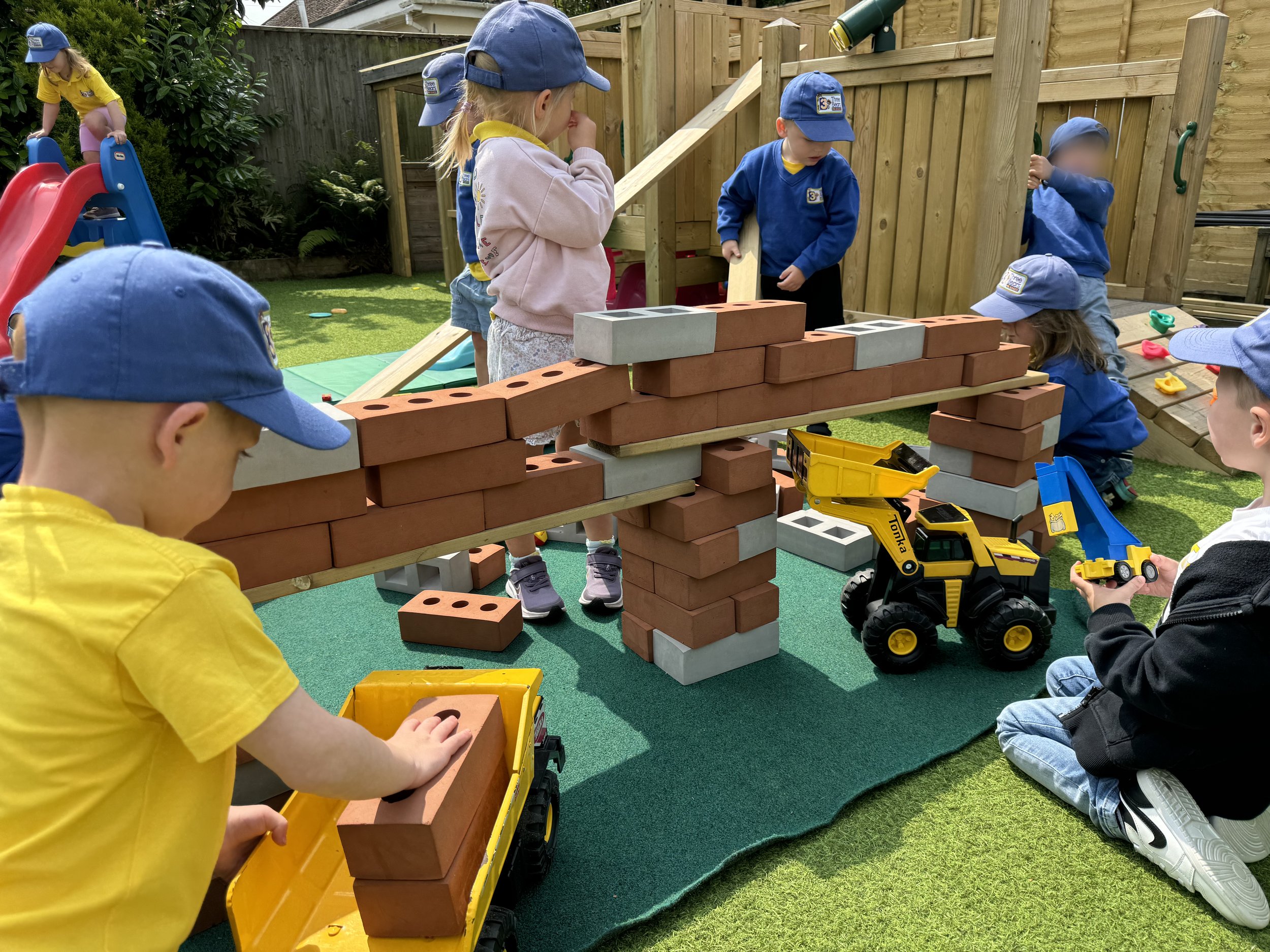 Children playing with large toy bricks and construction vehicles outdoors in a backyard playground, engaging in imaginative building activities.