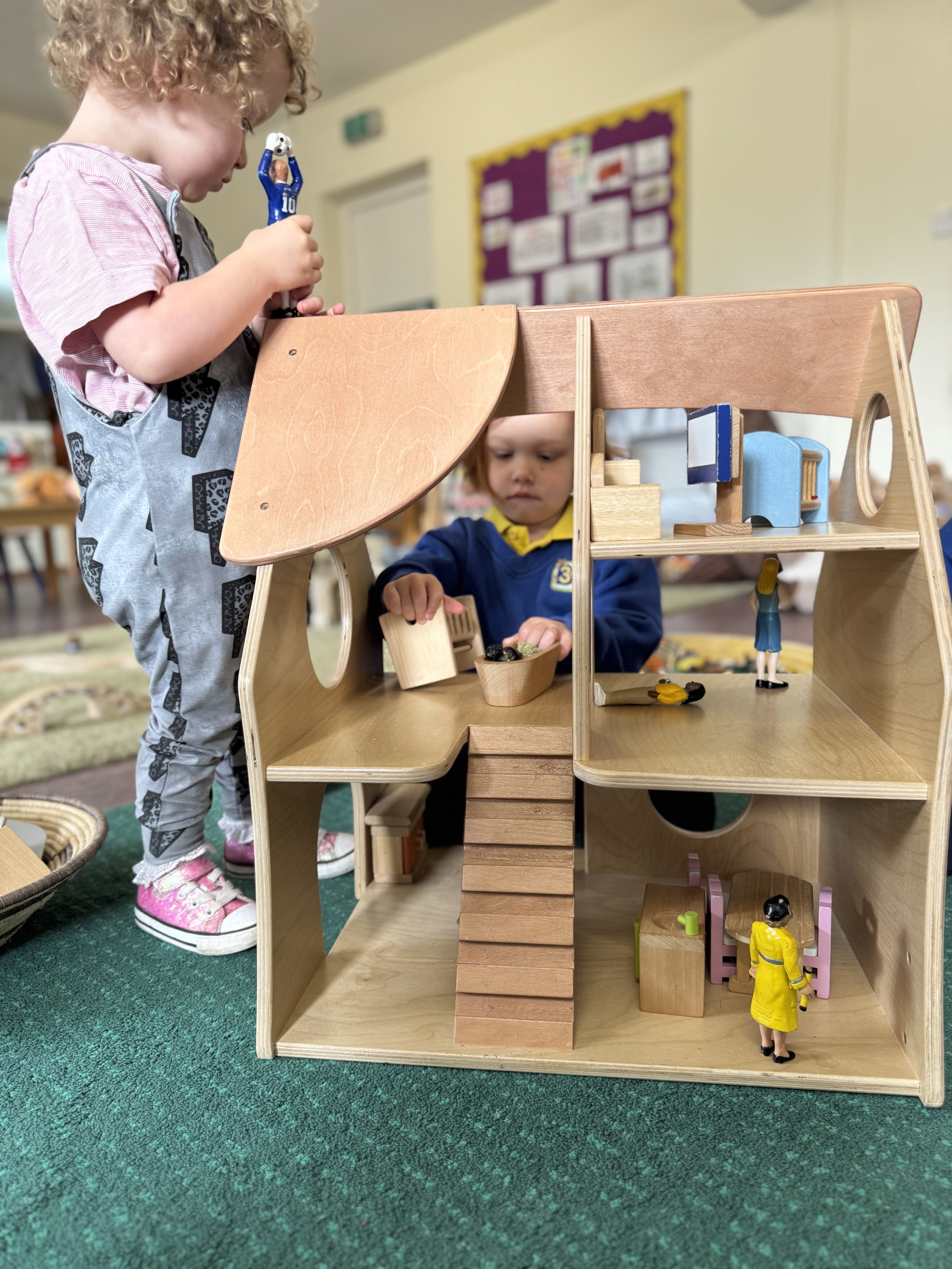 Two young children playing with a wooden dollhouse filled with small figurines in a classroom.