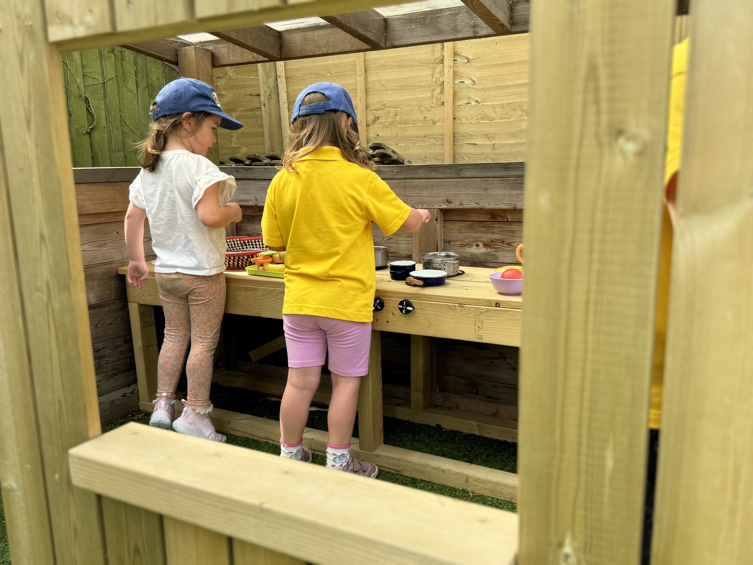 Two young girls playing in a wooden playhouse kitchen, wearing blue hats and casual clothing, pretending to cook with small pots and bowls.