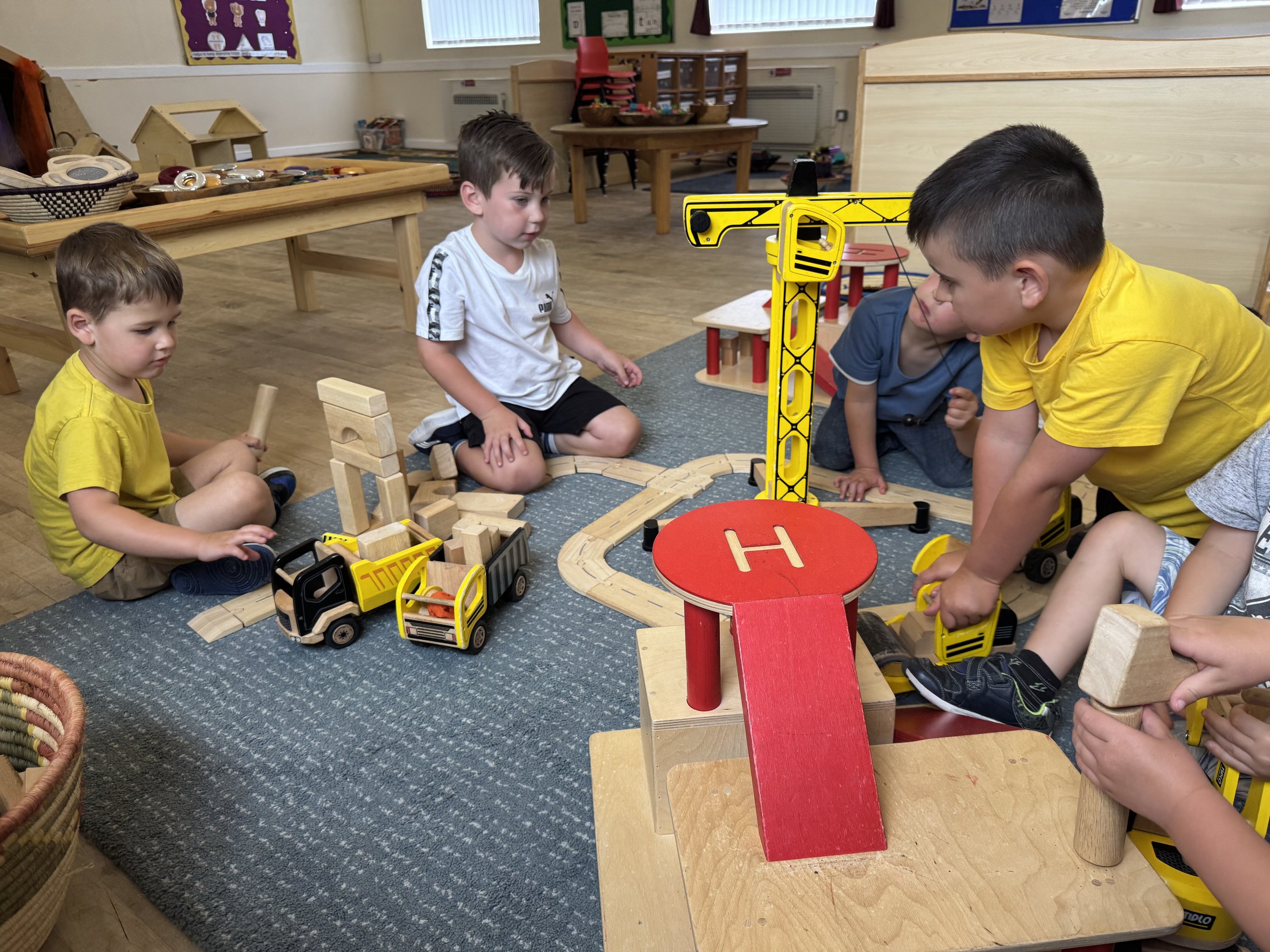 Four young children playing with toy construction vehicles and wooden blocks on a carpeted area in a classroom. Two children are seated on the carpet, one child is operating a yellow toy crane, and another is holding a wooden hammer. Various wooden b