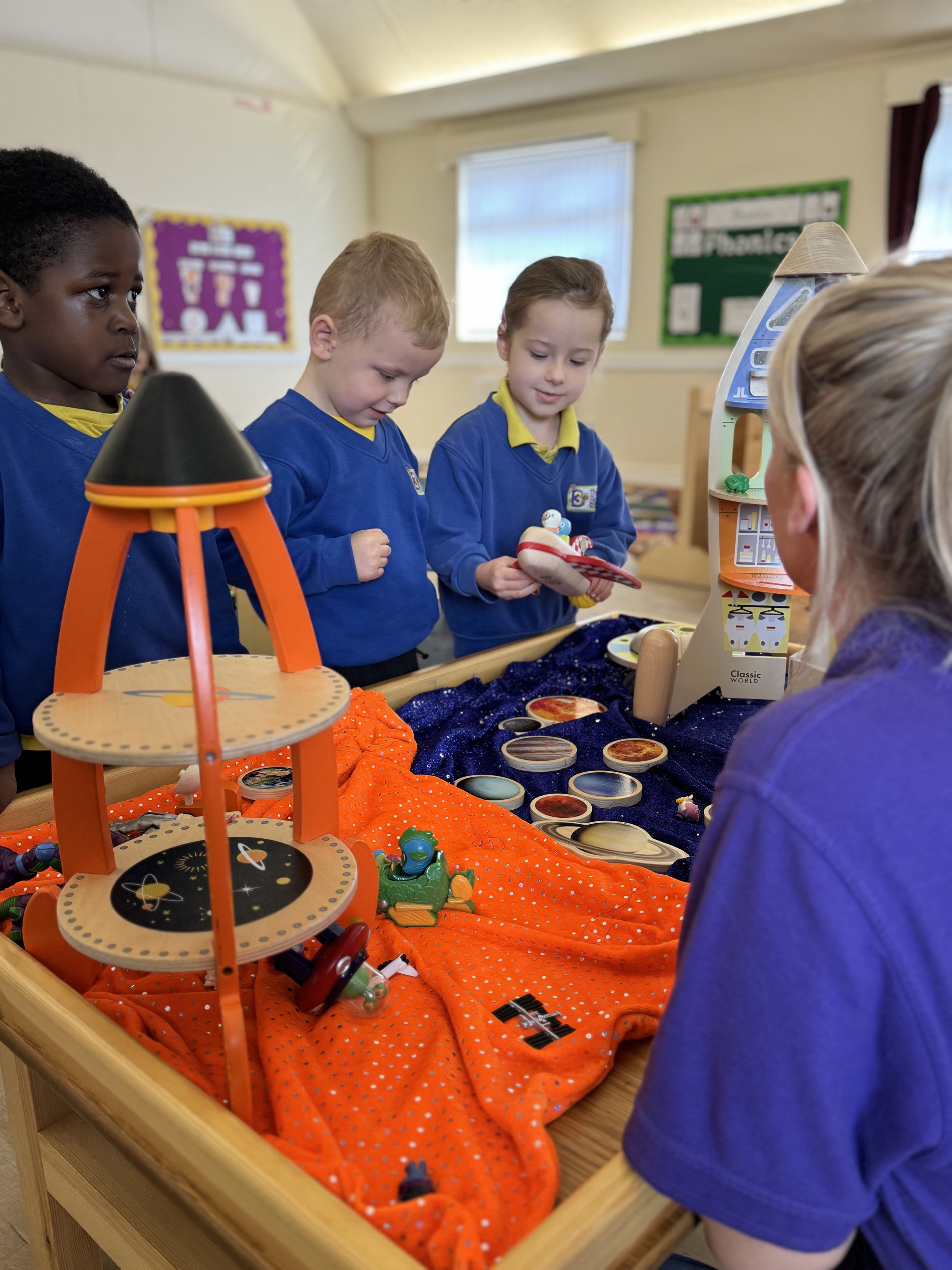Three children in blue school uniforms looking at space-themed toys and objects displayed on a table in a classroom.