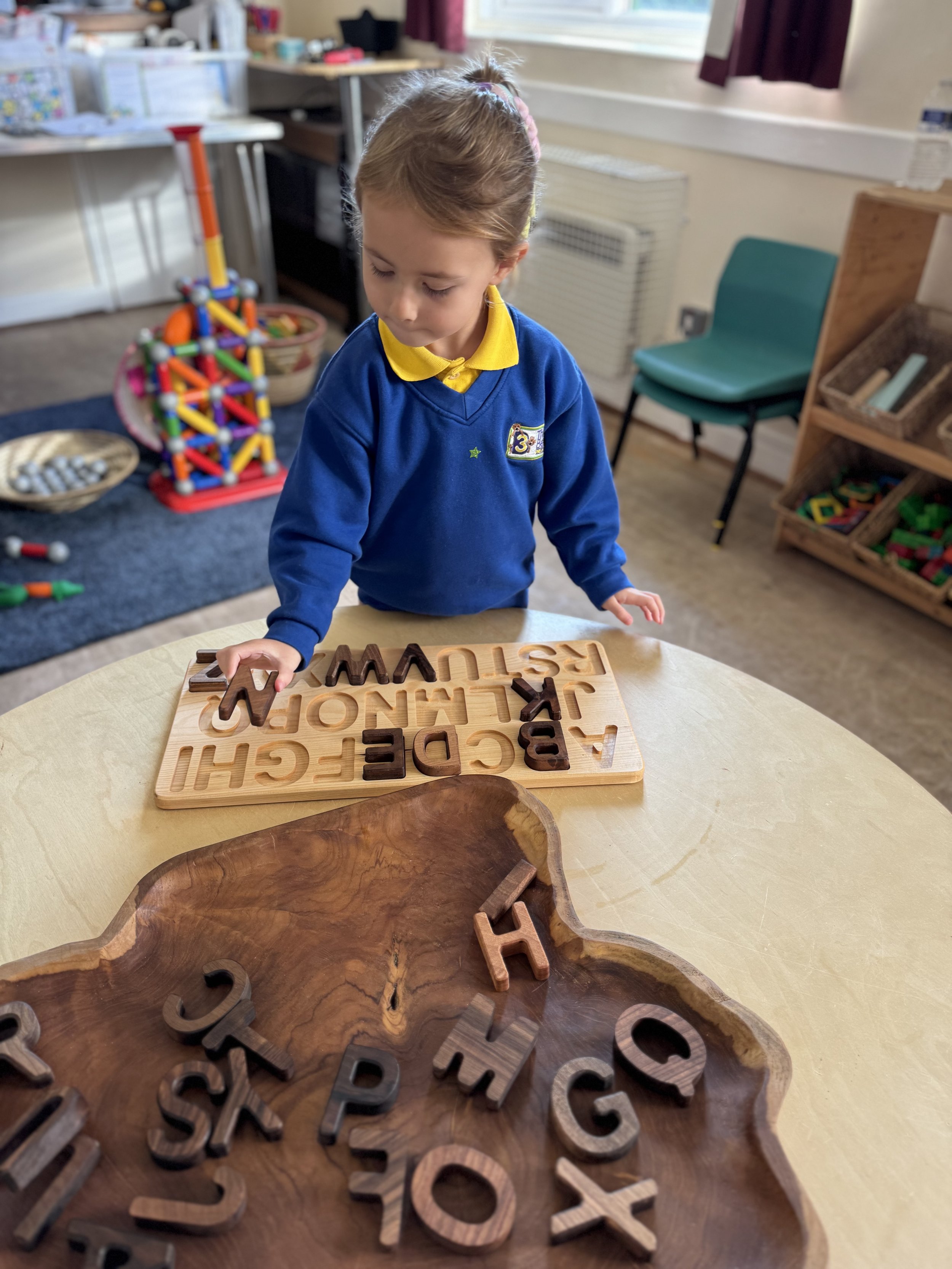A young girl in a blue school uniform with a yellow collar is playing with wooden alphabet puzzle pieces at a round table in a classroom.