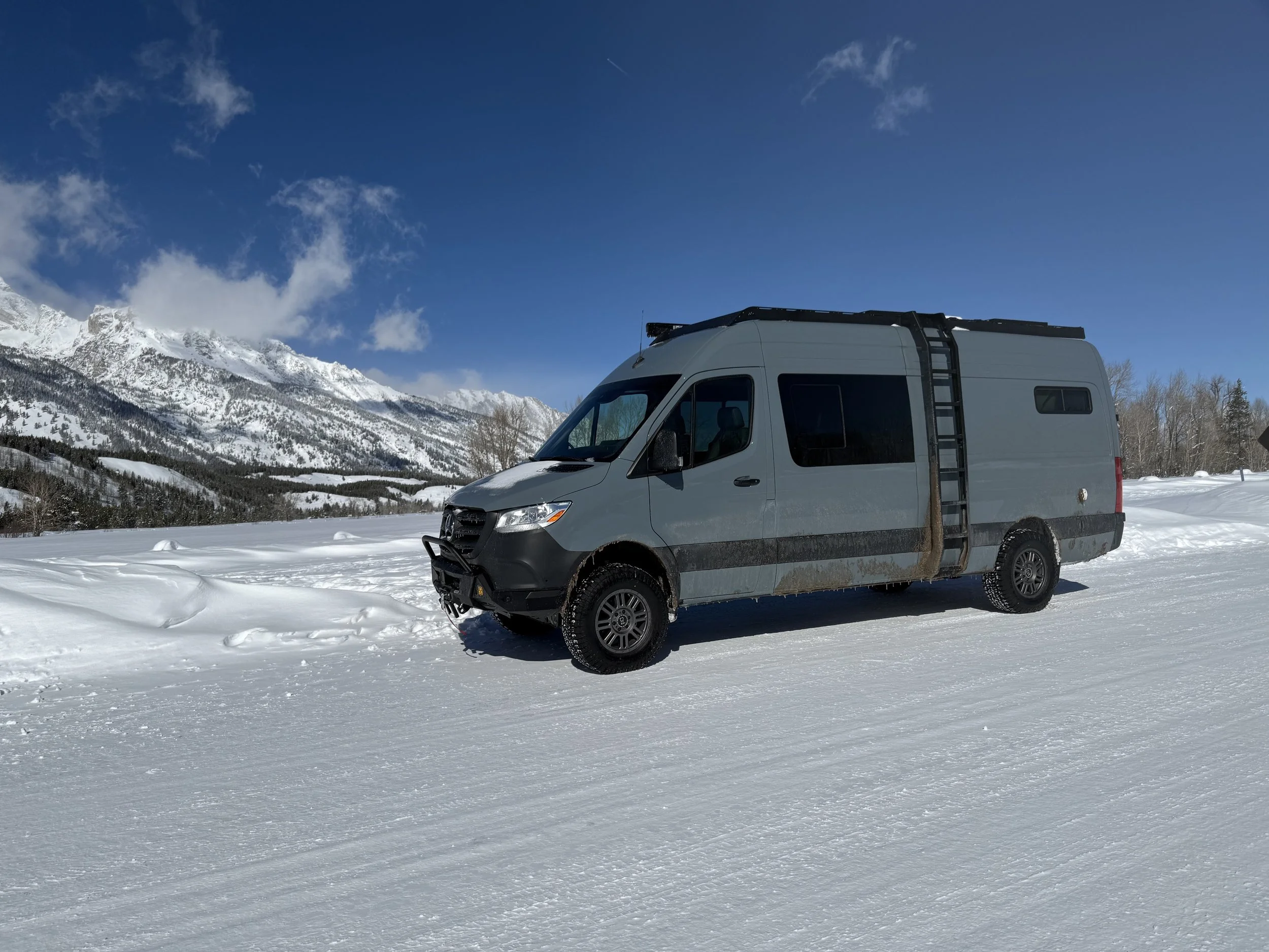 A gray camper van parked on a snowy landscape with mountains and a bright blue sky in the background.
