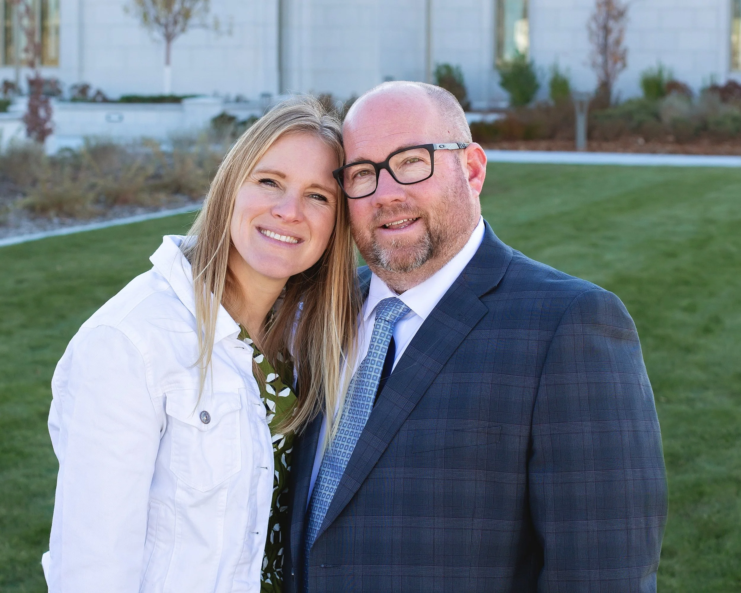 A smiling woman with blonde hair and a man with glasses and a beard, dressed in a suit, standing close together outdoors on a grassy area with trees and a building in the background.