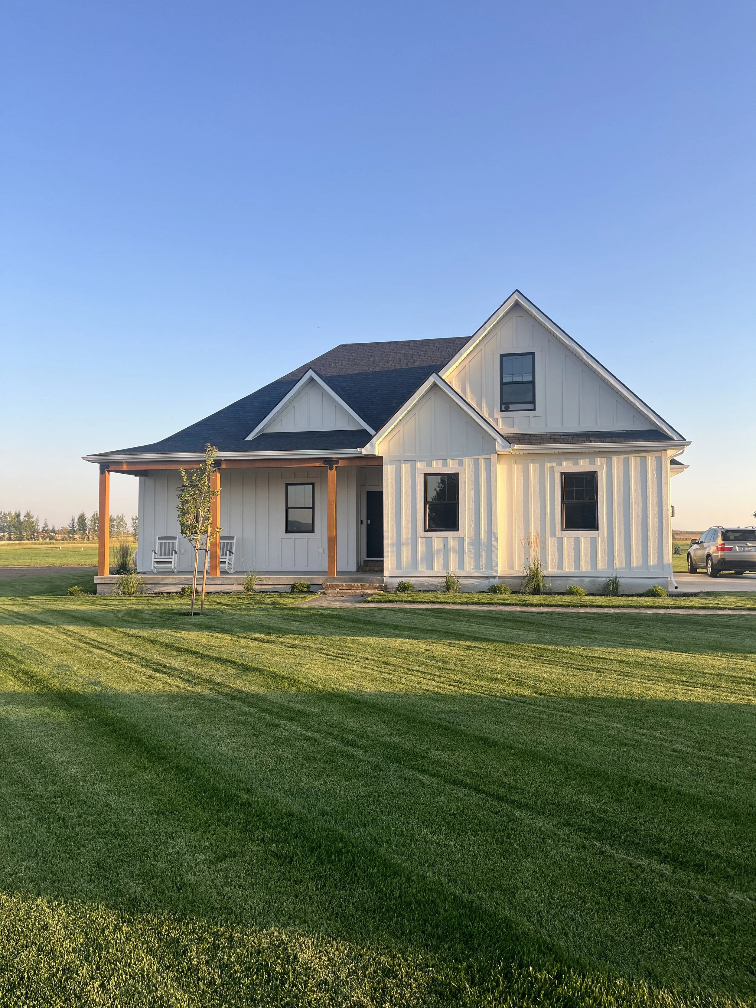 White house with black roof and wooden columns, front porch with benches, surrounded by green lawn and a few small trees, parked car on driveway, clear blue sky in the background.
