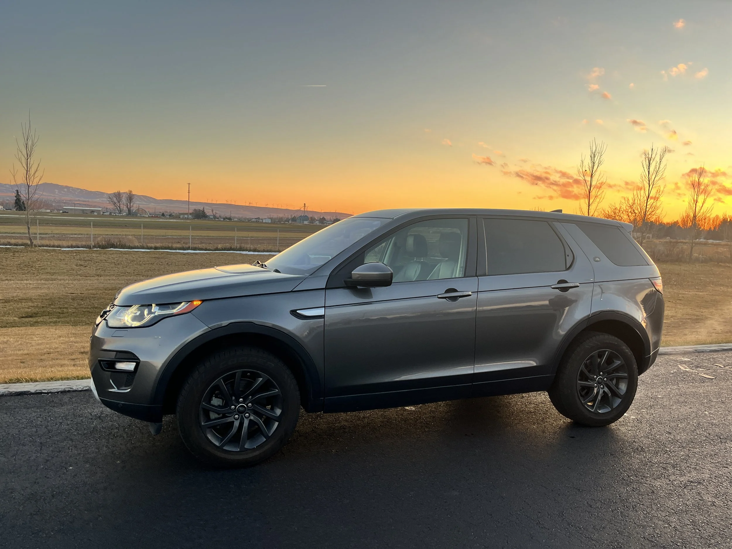 A metallic gray SUV parked on a paved surface during sunset with an open field and distant mountains in the background for rent in jackson hole