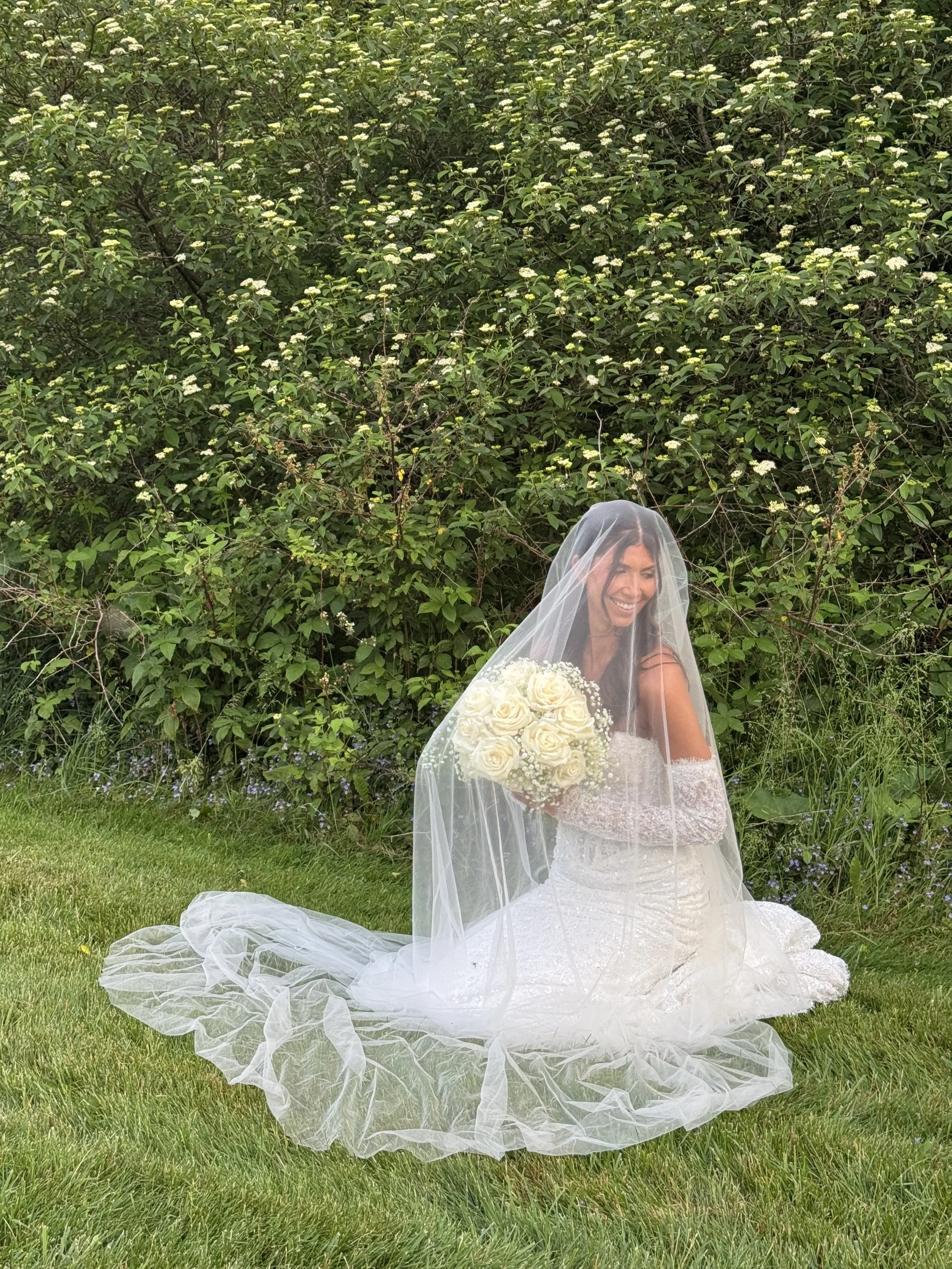 A bride dressed in a white wedding gown with lace details, holding a bouquet of white roses, sitting on the grass outdoors with a smile, partially covered by a sheer veil, in front of a green leafy bush.