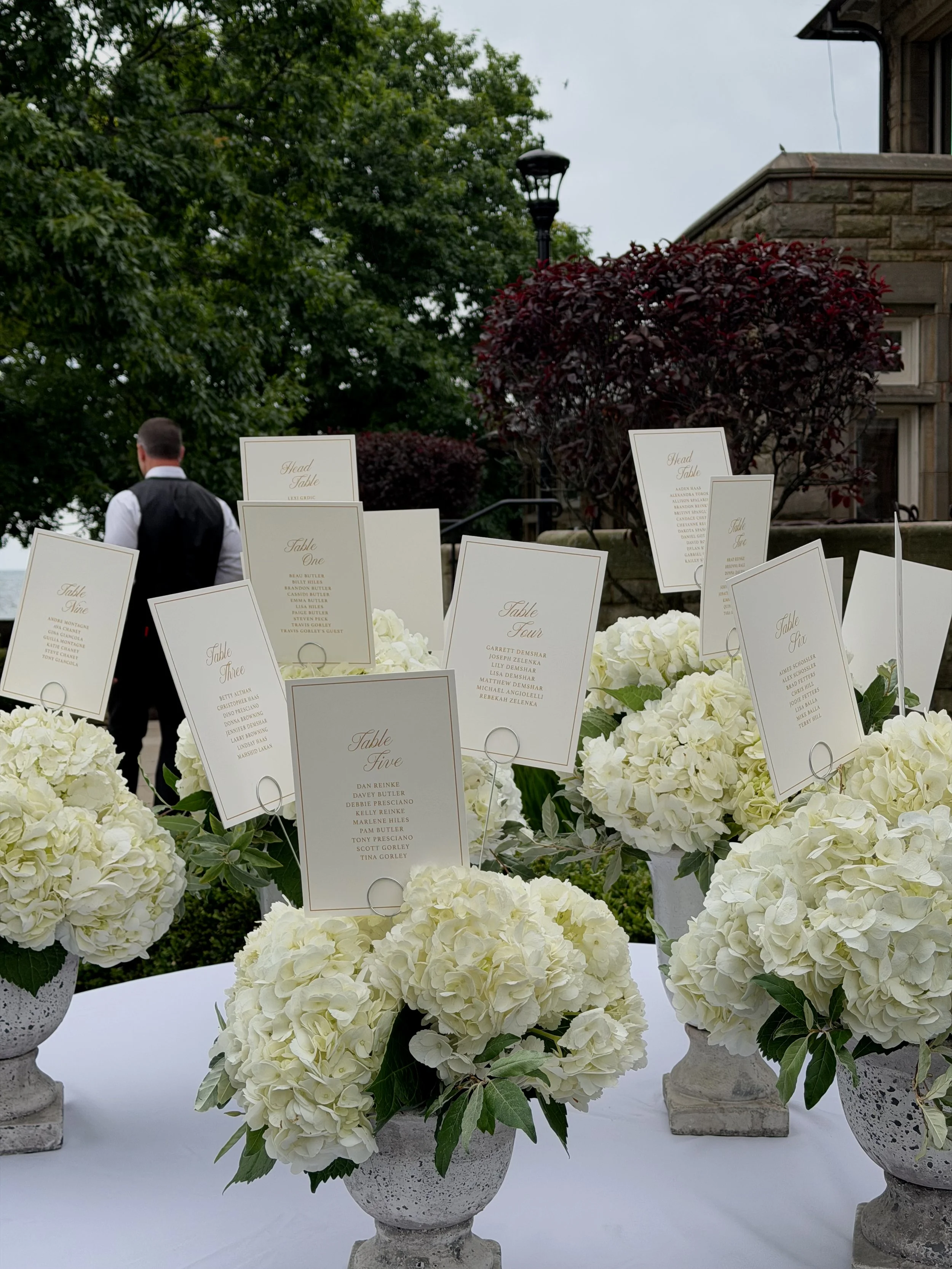 Long dining table with white tablecloth, place settings, and candles, set for an event, with black chairs and floral decorations. Visible three people standing near the table. Checkered black and white floor.