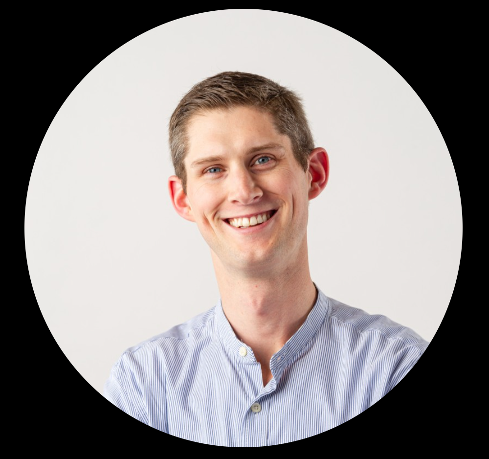 A young man with short brown hair and blue eyes, smiling, wearing a light blue striped button-up shirt on a plain white background.