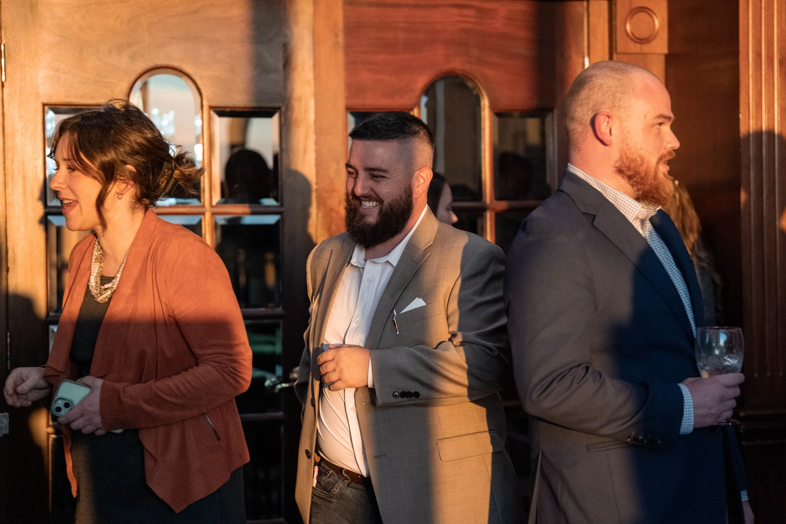 Three people standing in a line at a social event, smiling and engaging, with a wooden interior background.