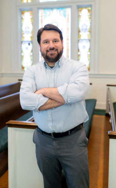 A man with dark hair and a beard, wearing a light blue shirt and gray pants, standing with arms crossed inside a church with stained glass windows.