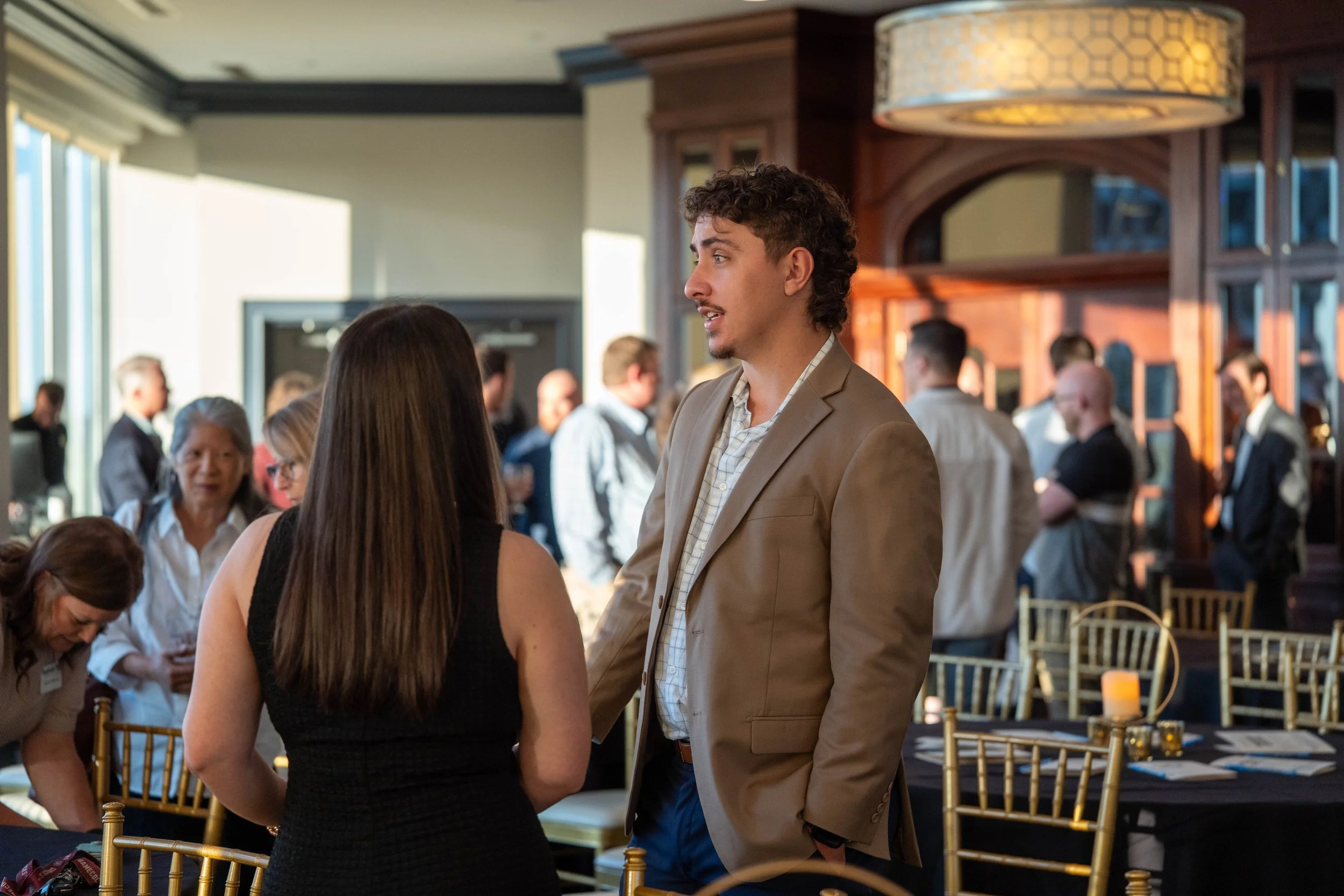 A man in a tan blazer and a woman with long brown hair in a black dress are talking at a social event in a restaurant or banquet hall. People are mingling in the background, and there are tables with chairs and candles.