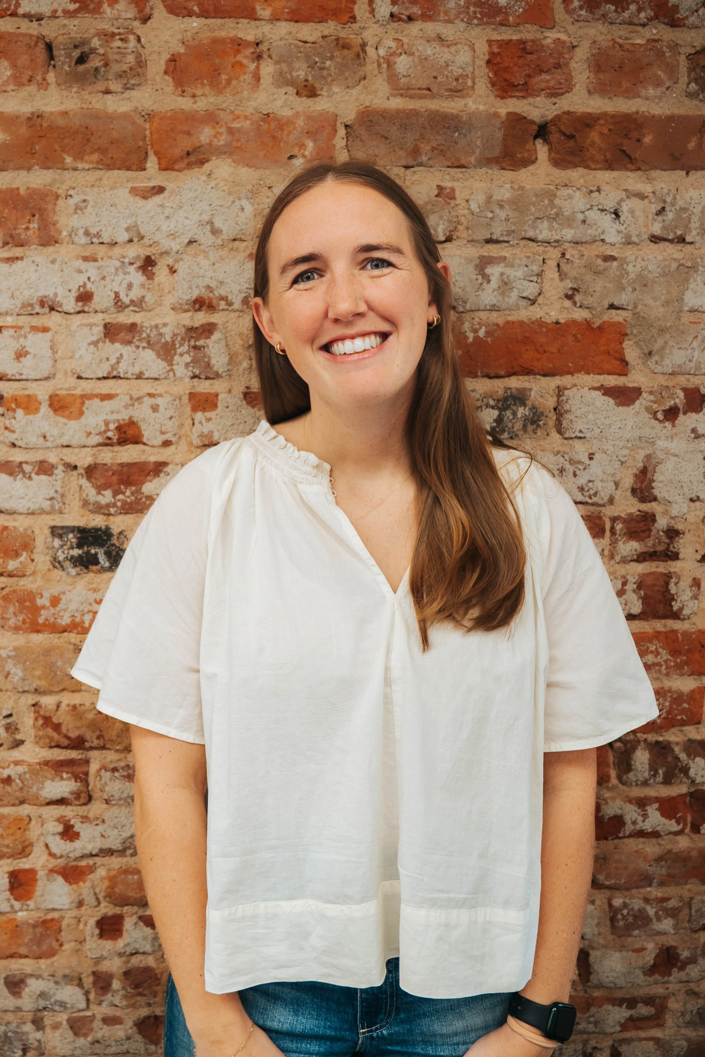 A woman with long brown hair wearing a white blouse, gold earrings, and a black wristwatch, smiling in front of a brick wall.