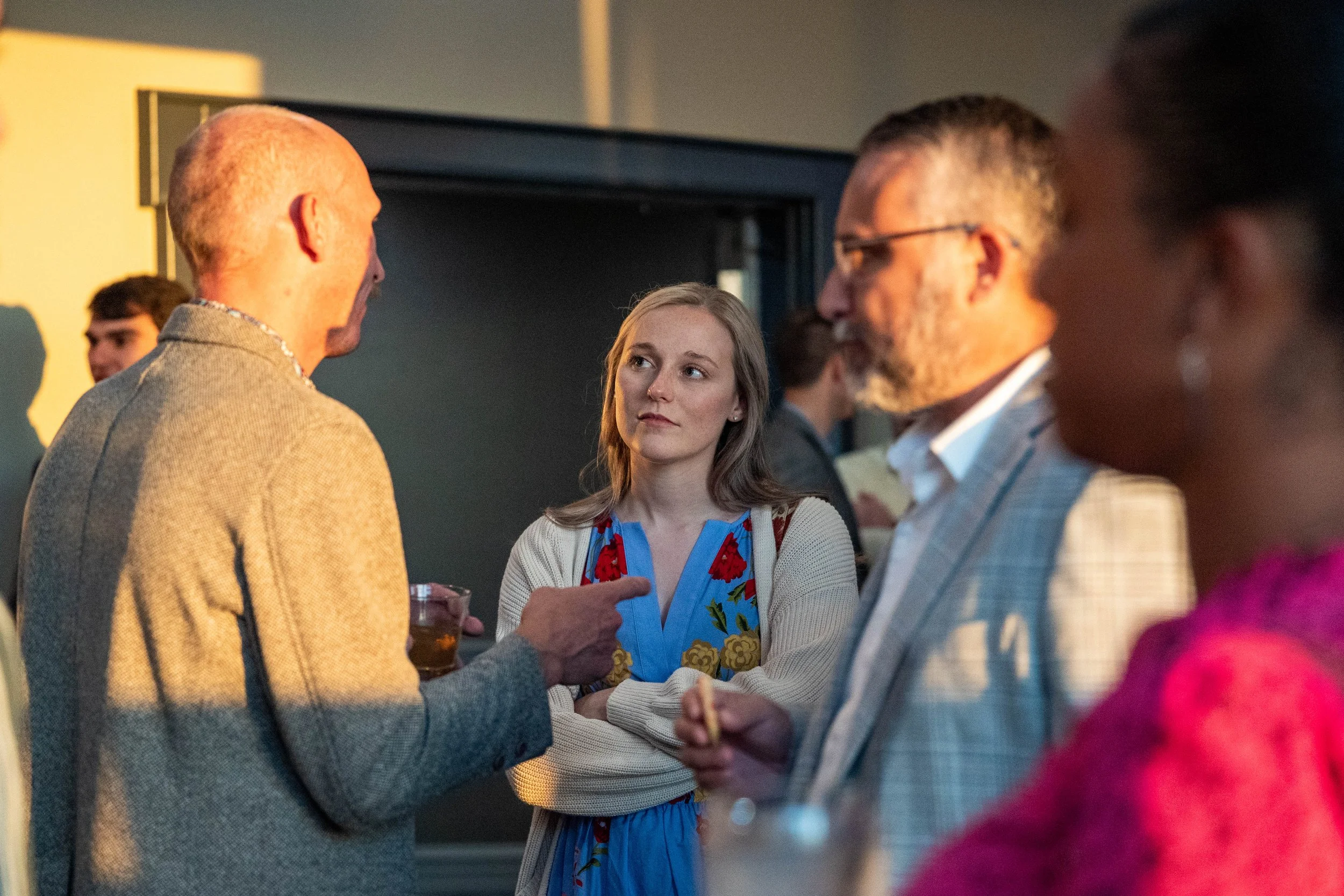 Group of four adults talking at a social event, with a woman in a colorful dress and cardigan listening to a man with a glass of drink, in a warmly lit indoor setting.
