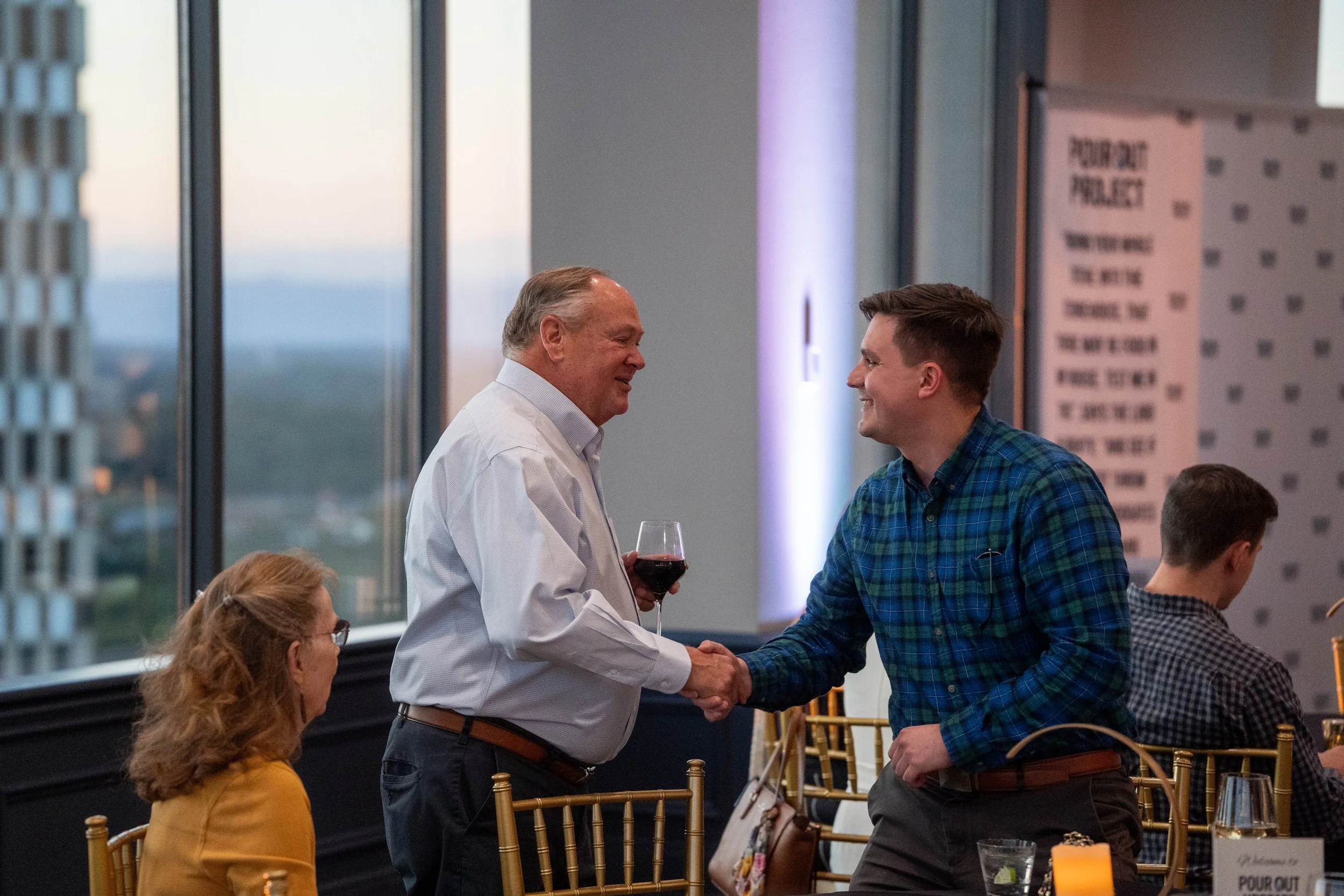 Two men shaking hands in a restaurant, with a woman watching. One man holds a glass of red wine. The restaurant has large windows showing a cityscape at sunset.