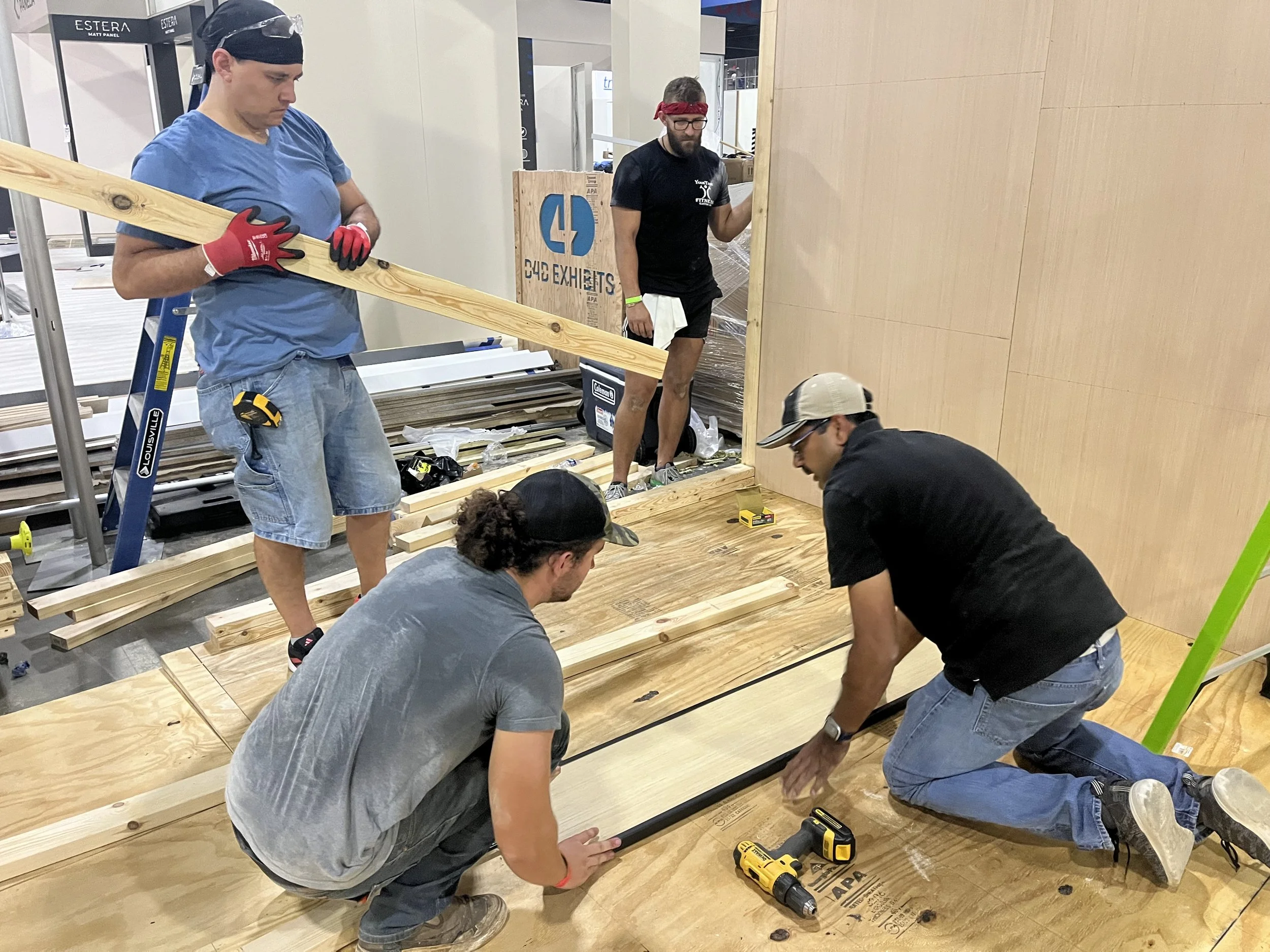 A group of men assembling wooden furniture or framing, with tools and wooden planks on the floor, in an indoor setting.