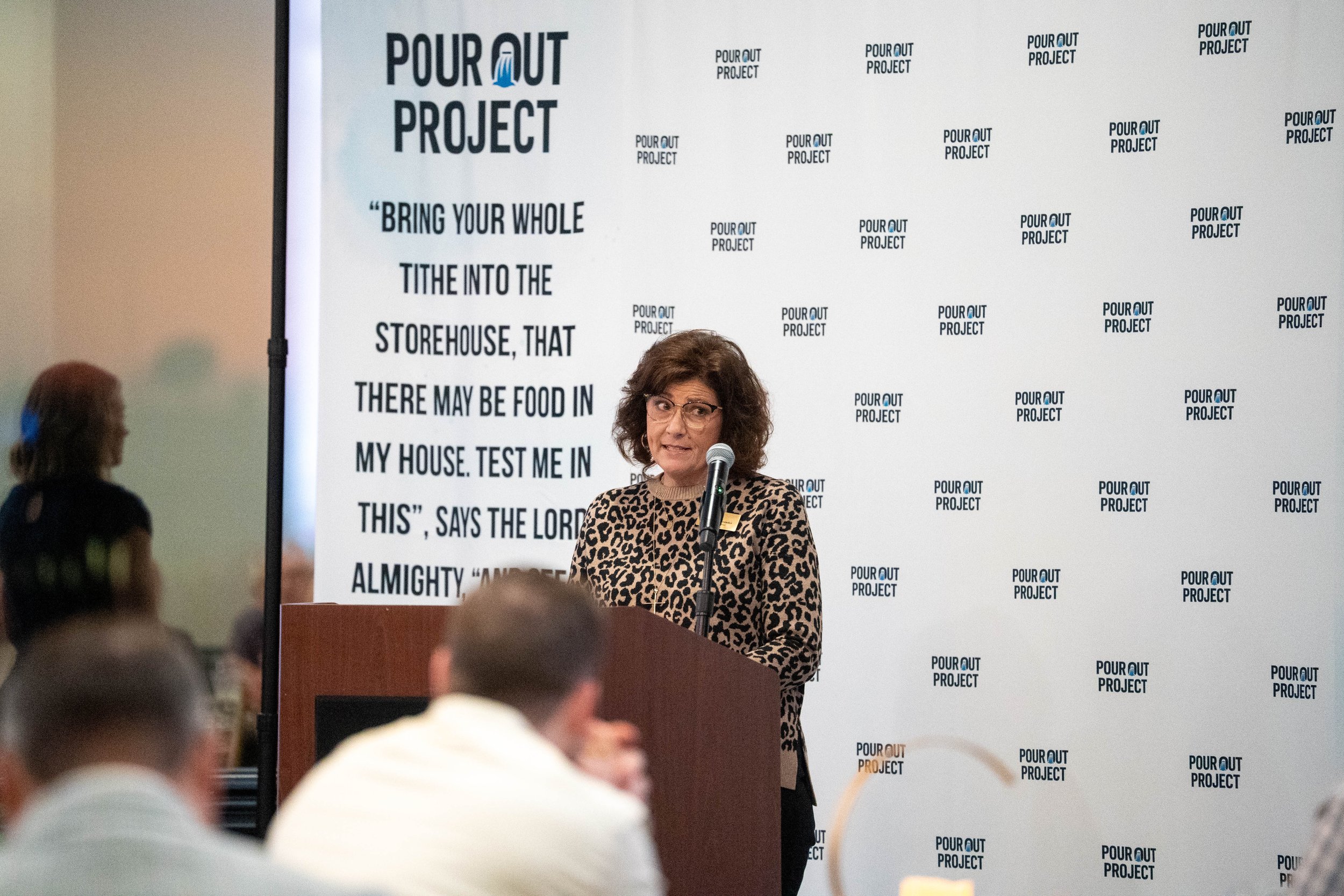 A woman with curly hair and glasses speaks at a podium during a presentation. Behind her is a large screen with the words "Pour Out Project" and a Bible quote about bringing food into the storehouse.
