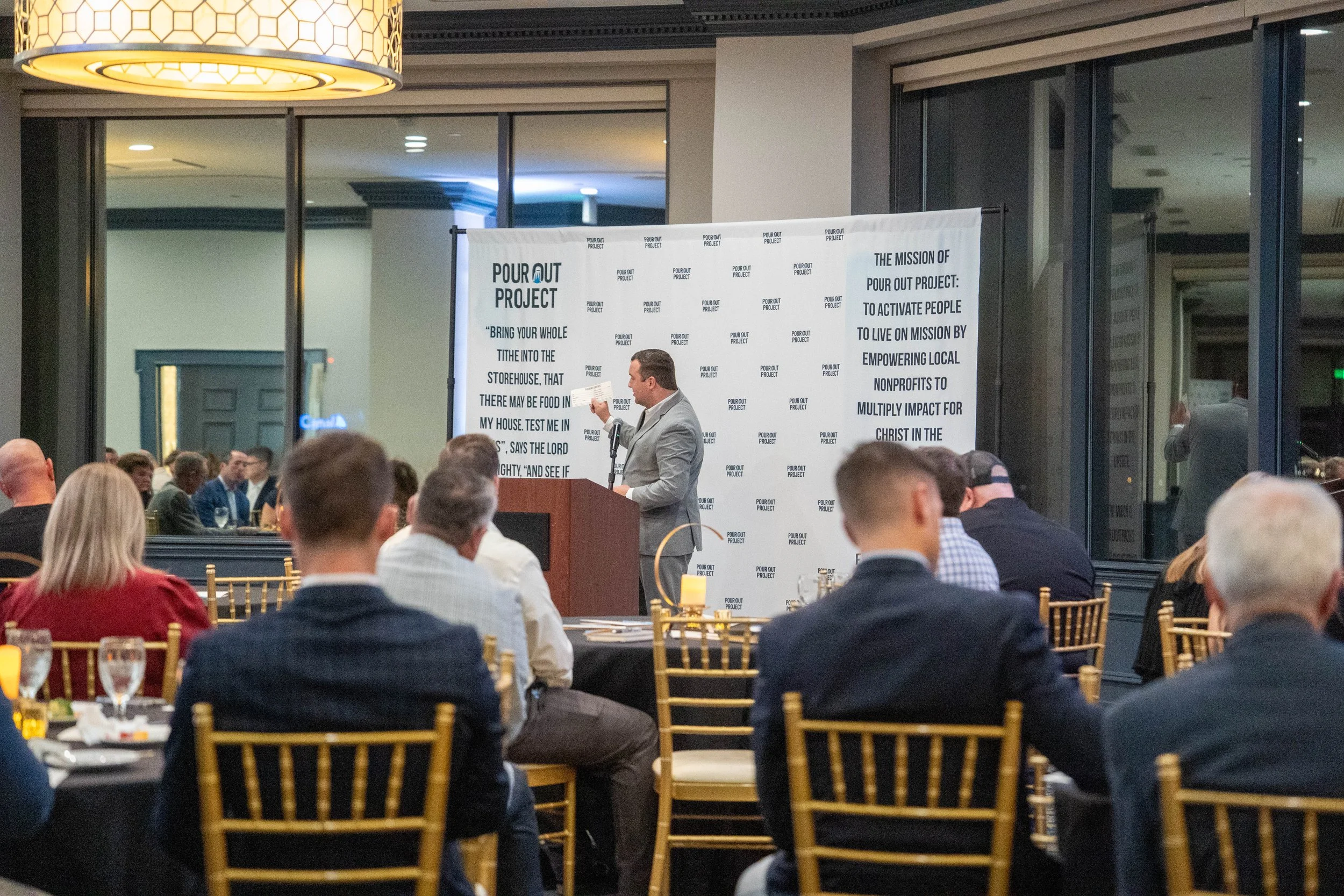 A man in a gray suit giving a presentation at a podium during a formal event or conference. A large white banner with text about a project is behind him, and audience members are seated at tables with black tablecloths and gold chairs.