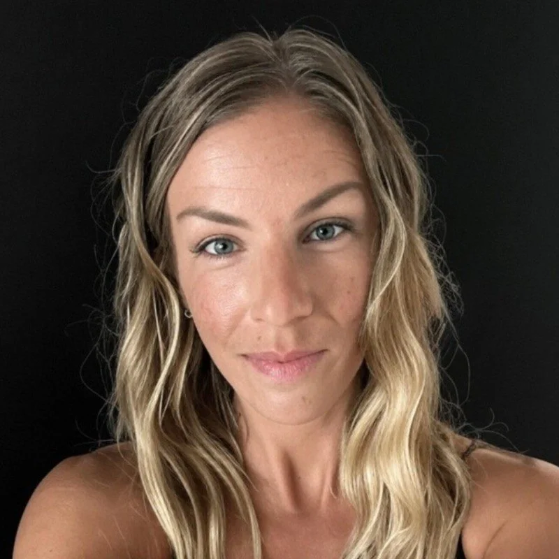 A woman with blonde, wavy hair and blue eyes smiling softly against a black background.