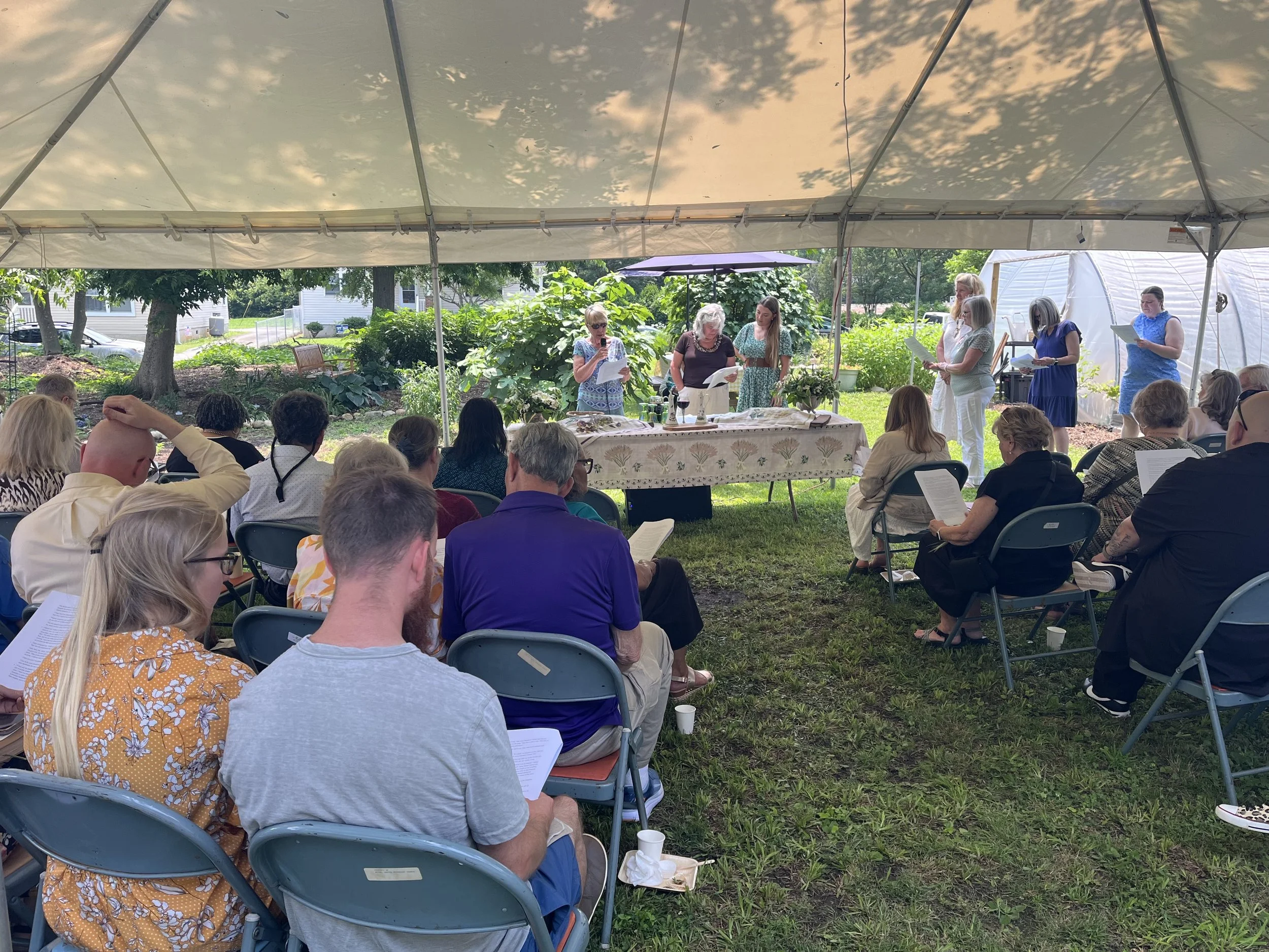 A group of people attending an outdoor gathering in a garden under a large canopy, with some reading from papers and a table with items in front of them, surrounded by green trees and plants.